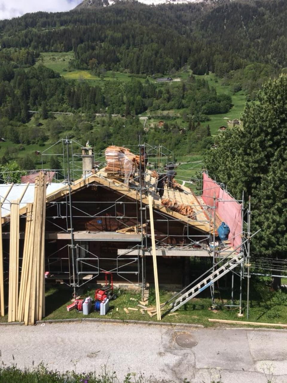Maison en construction ; des échafaudages entourent le toit, laissant apparaître la charpente en bois et la cheminée. Montagne en arrière-plan.
