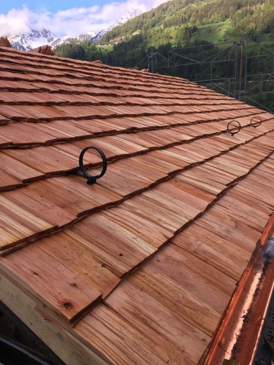 Toit en bardeaux de cèdre avec des anneaux métalliques noirs sur fond de montagne boisée.
