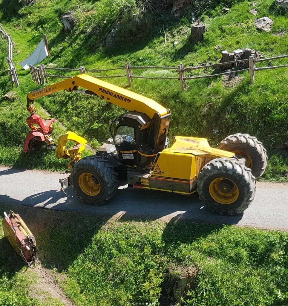Engin forestier jaune sur une route dans un environnement verdoyant et vallonné, avec un bras de grappin rouge.