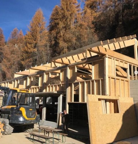 Chantier de construction d'un bâtiment à ossature bois, avec une pelleteuse jaune au premier plan. Arbres aux feuilles orangées en arrière-plan.