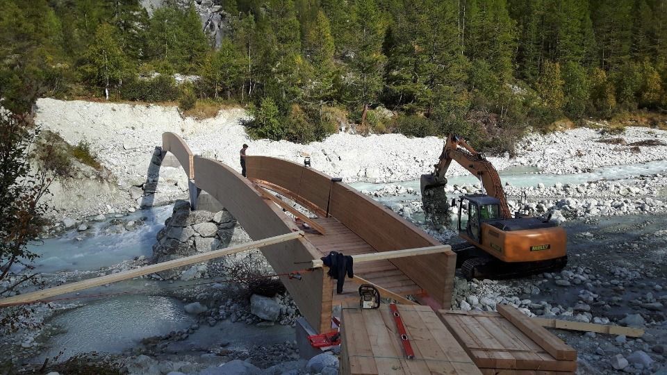 Un pont en construction au-dessus d'un lit de rivière rocailleux, avec une excavatrice, du bois et une forêt en arrière-plan.