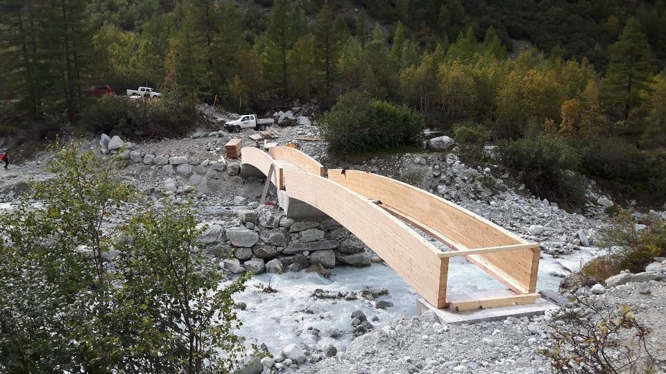 Pont en bois en construction au-dessus d'un lit de rivière rocailleux, entouré d'arbres.