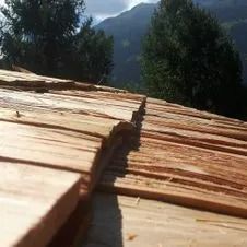 Maison en construction ; des échafaudages entourent le toit, laissant apparaître la charpente en bois et la cheminée. Montagne en arrière-plan.