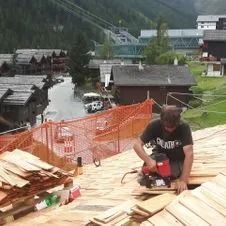 Construction d'une charpente de toit : une grue soulève une poutre en bois. Un ouvrier se tient sur le toit.
