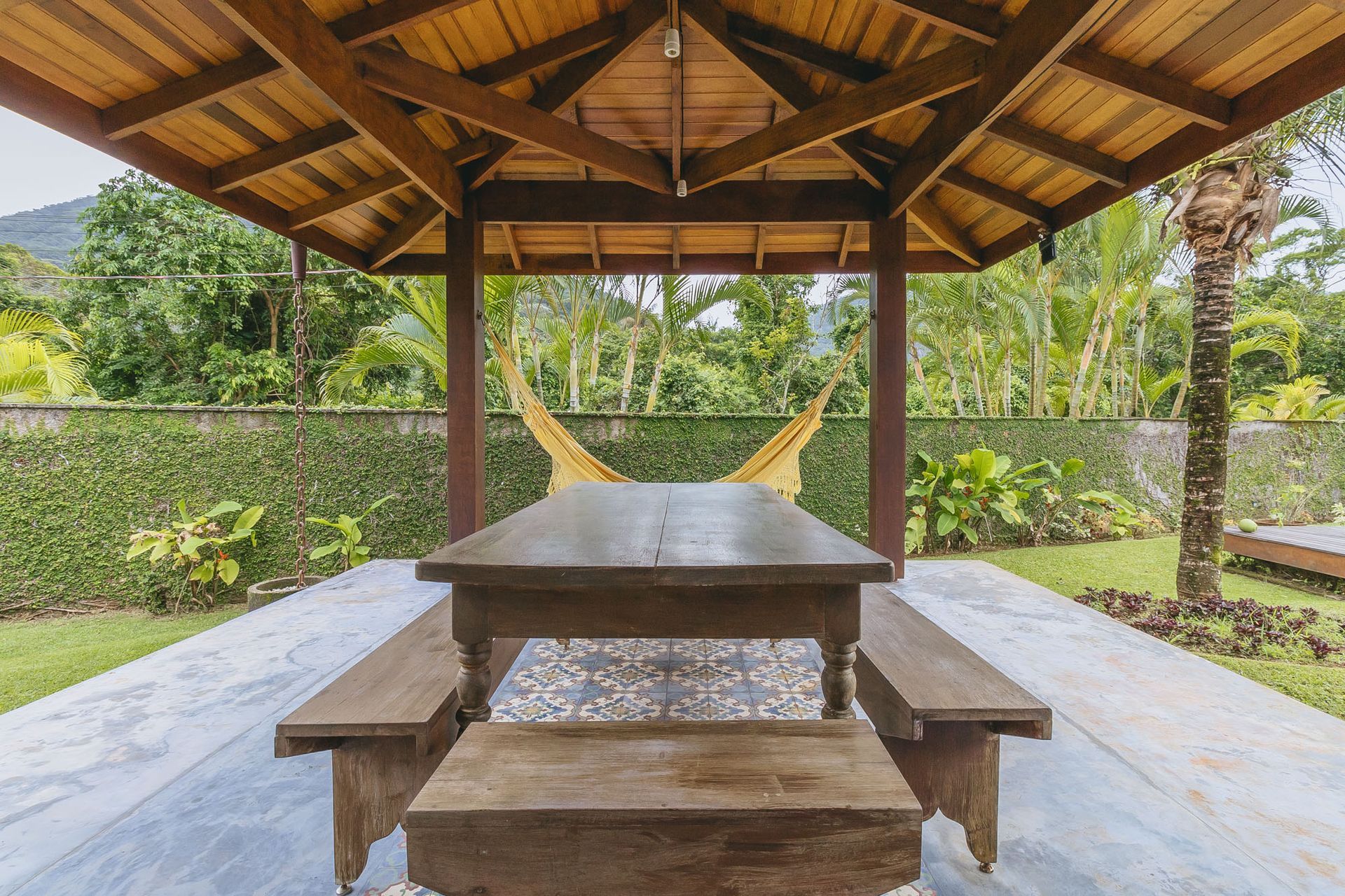 Gazebo de madeira com mesa e bancos com vista para um quintal verdejante.