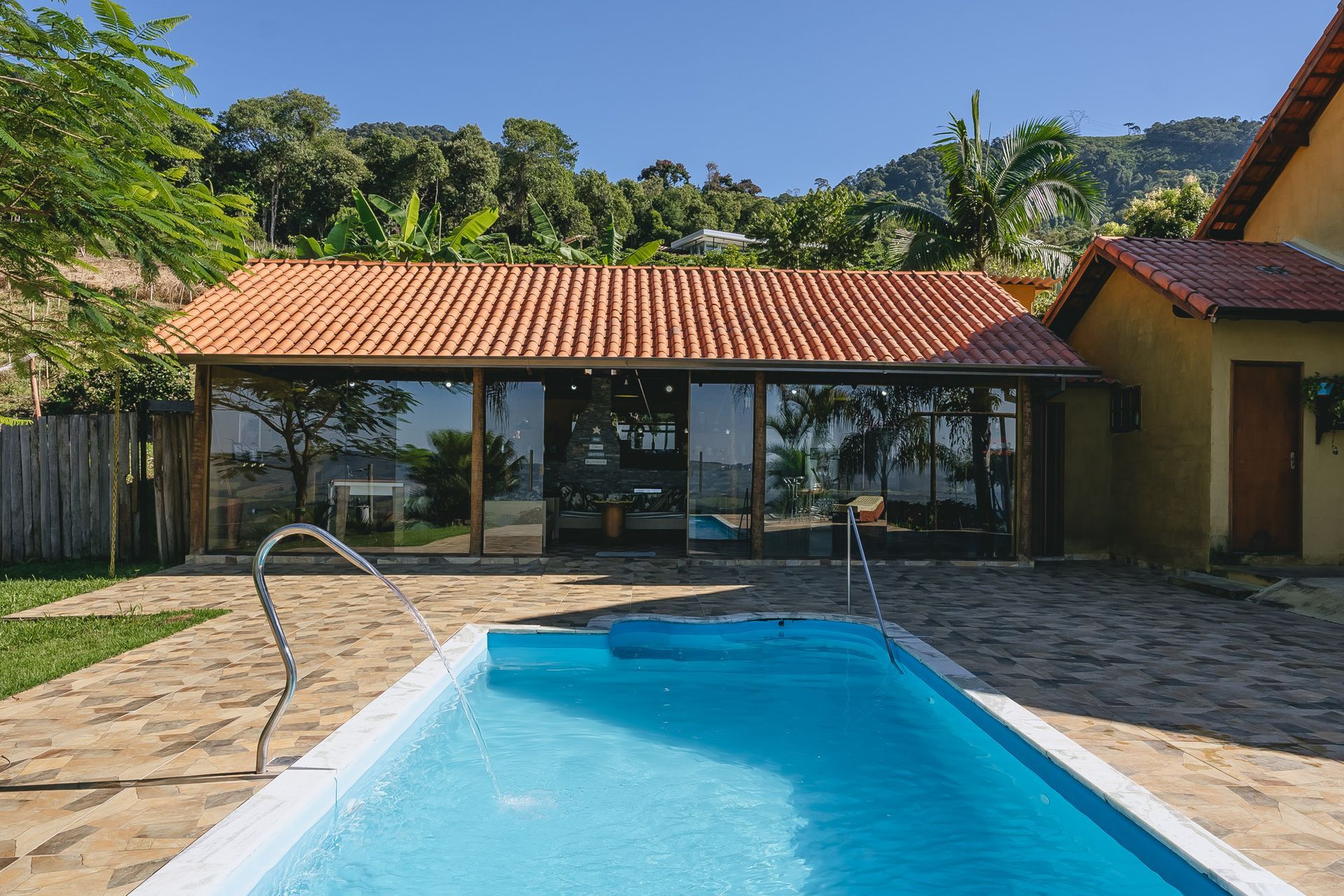 Piscina em frente a um edifício com paredes de vidro e telhado de telhas, em um cenário verdejante.