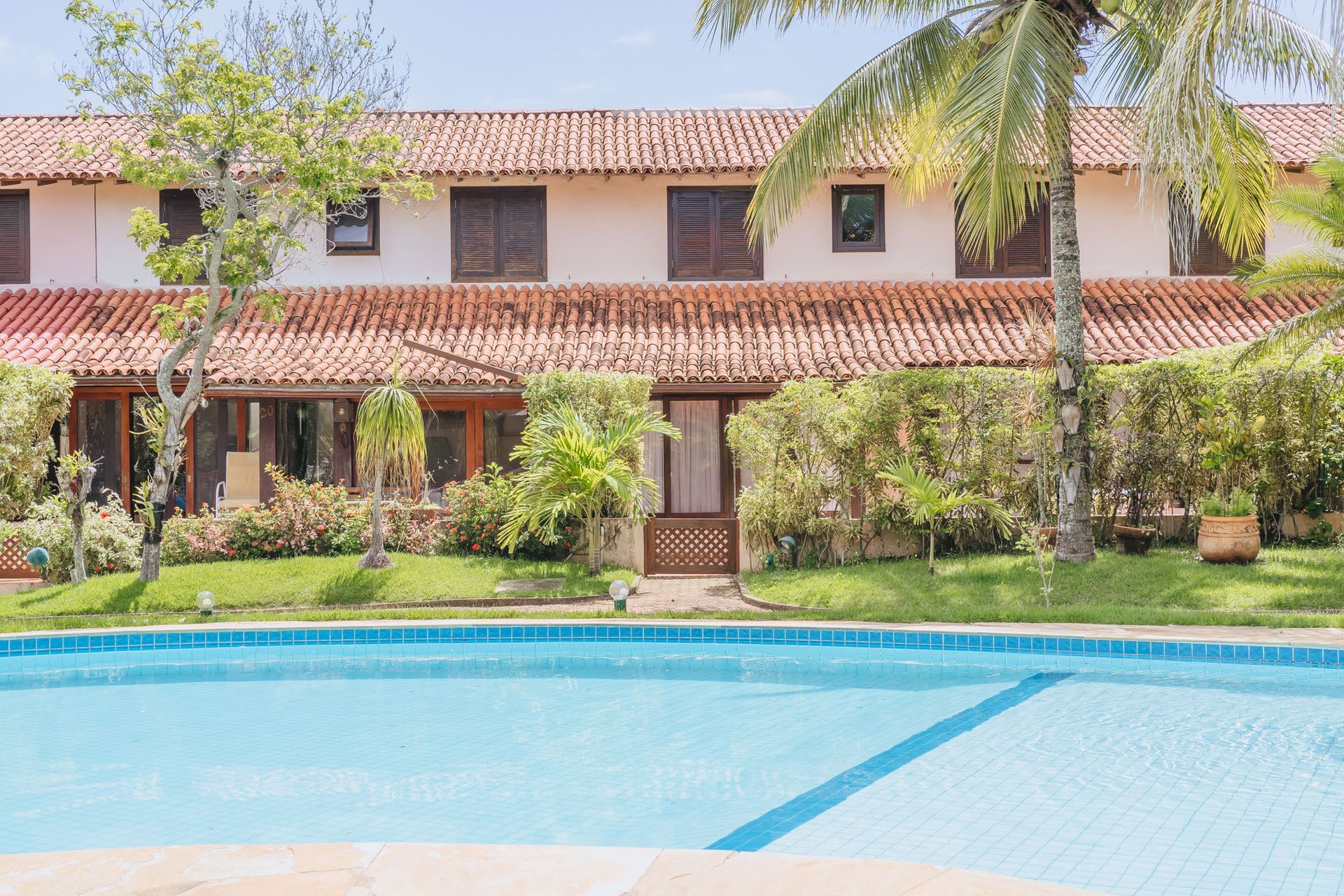 Um edifício de dois andares com piscina, cercado por vegetação e palmeiras.