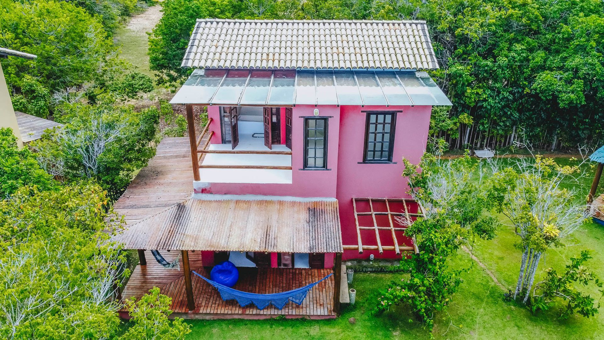 Casa rosa de dois andares com varanda com telhado de palha, sacada e árvores verdes exuberantes.