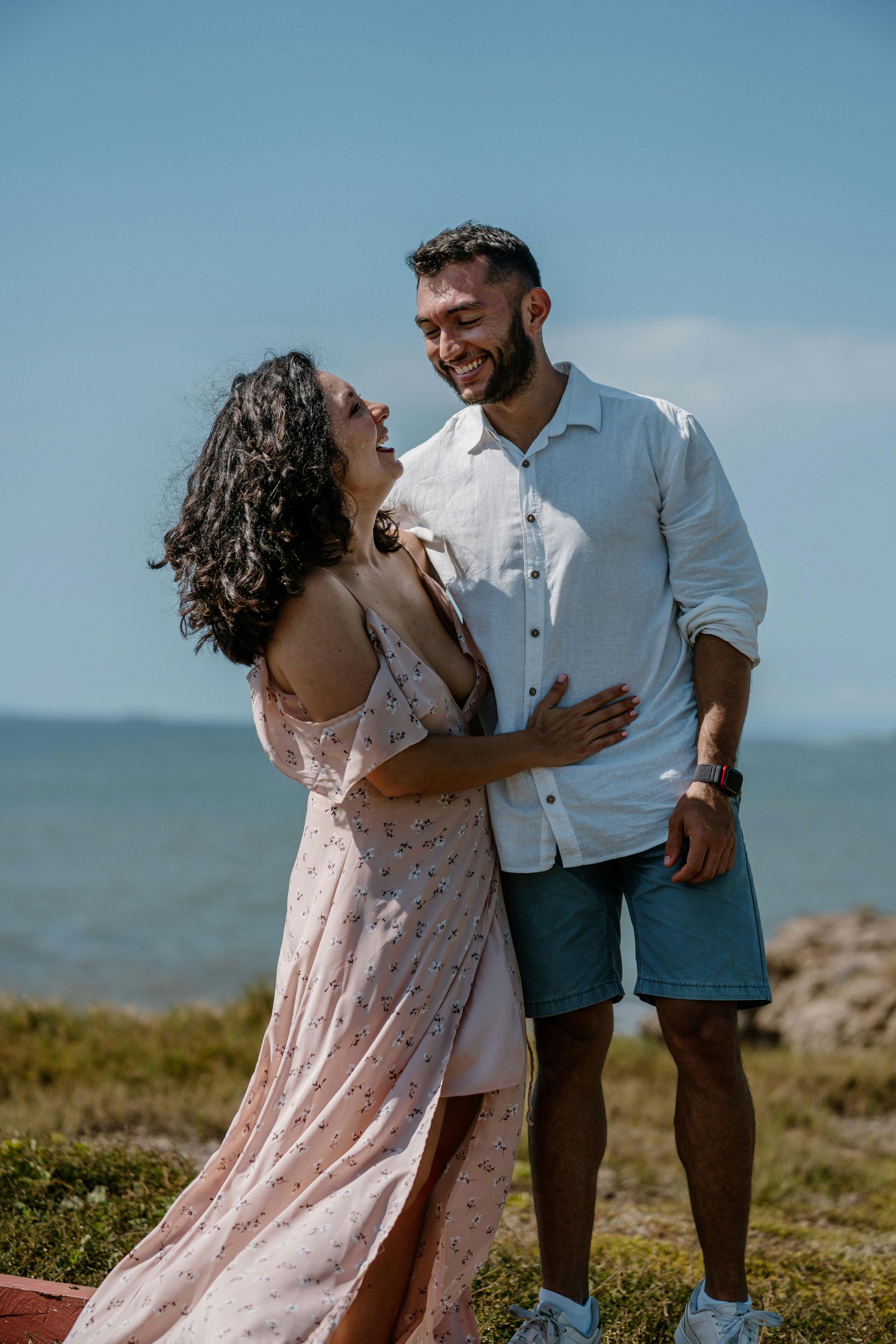 Casal rindo, abraçado à beira-mar. Mulher de vestido rosa, homem de camisa branca e shorts.