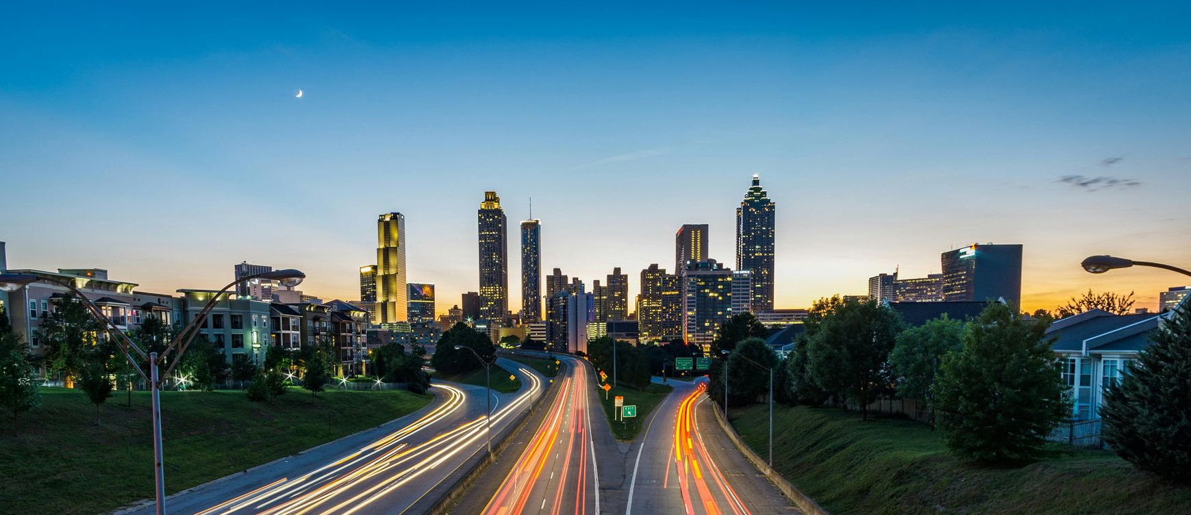Vista do horizonte de Atlanta ao entardecer, com rastros de luz de longa exposição provenientes do tráfego em uma rodovia em primeiro plano.
