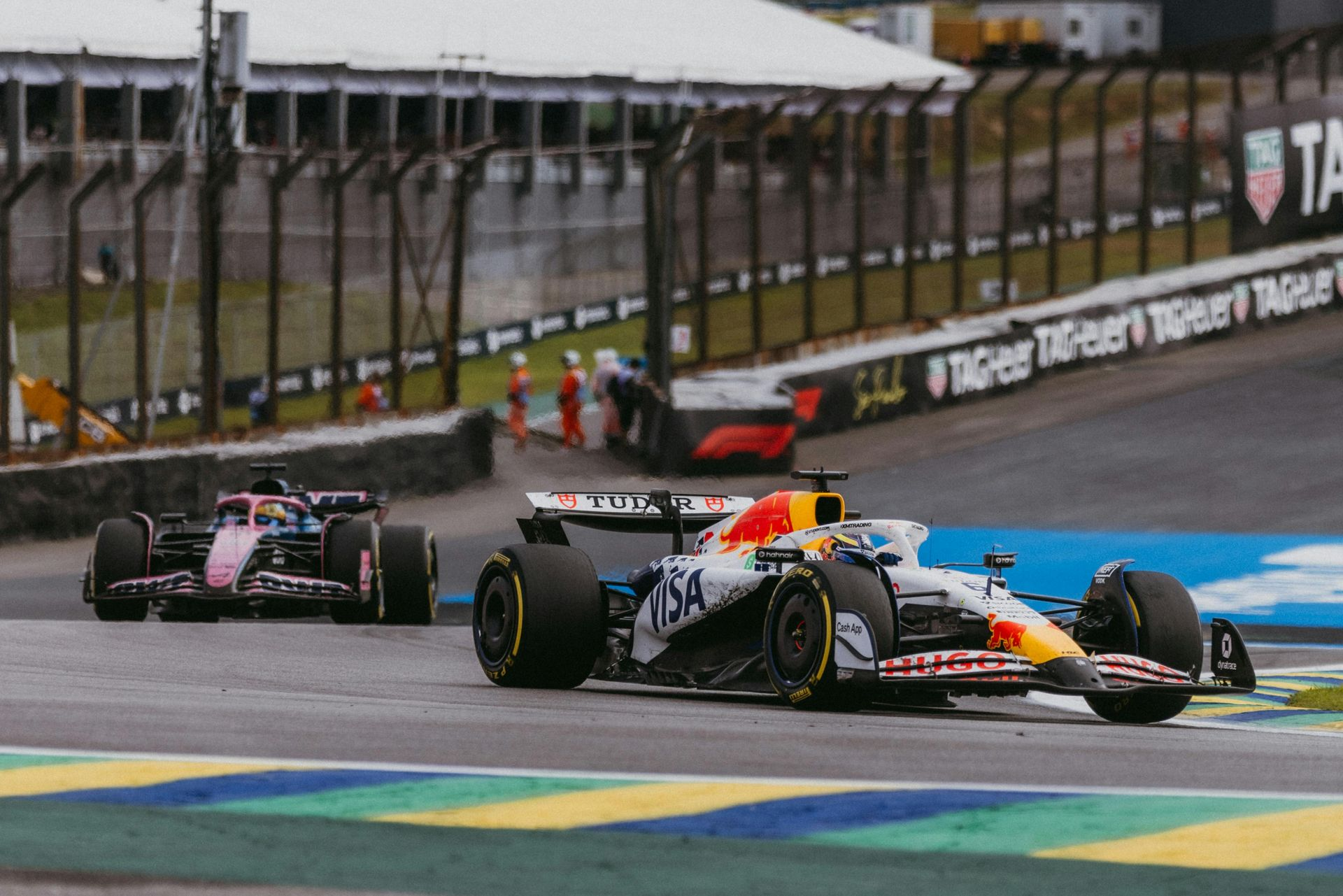 Dois carros de Fórmula 1 aceleram em uma pista curva em um circuito com a sinalização da TAG Heuer ao fundo.