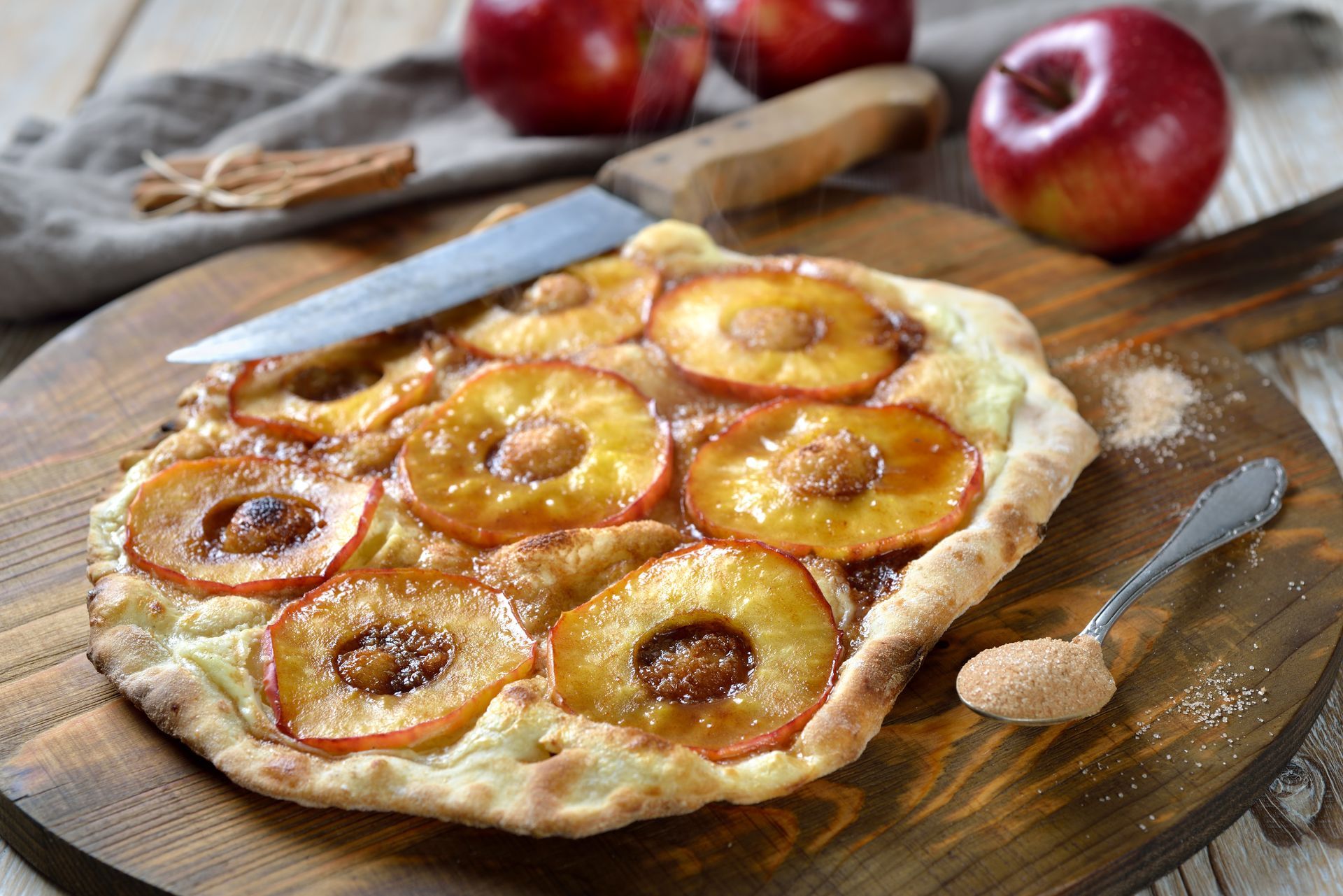 Tarte aux pommes sur une planche en bois, garnie de tranches de pommes glacées. Sucre à la cannelle parsemé à proximité.