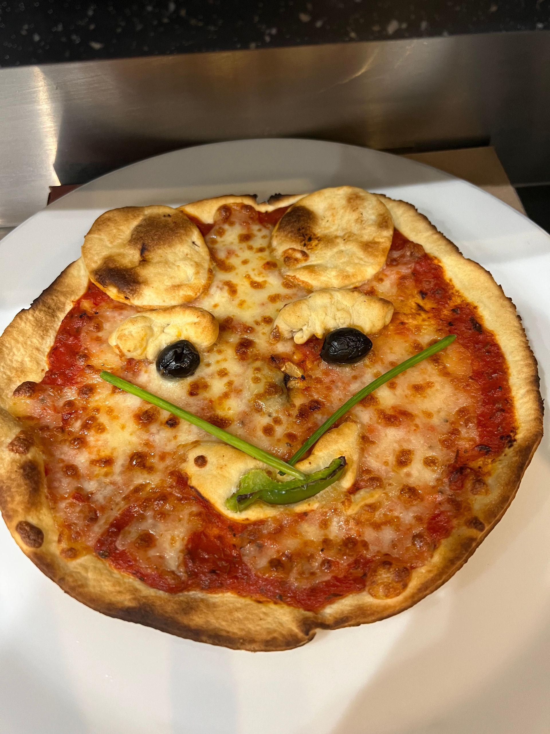 Une pizza souriante avec du fromage, de la sauce tomate, des olives et du poivron vert sur une assiette blanche.