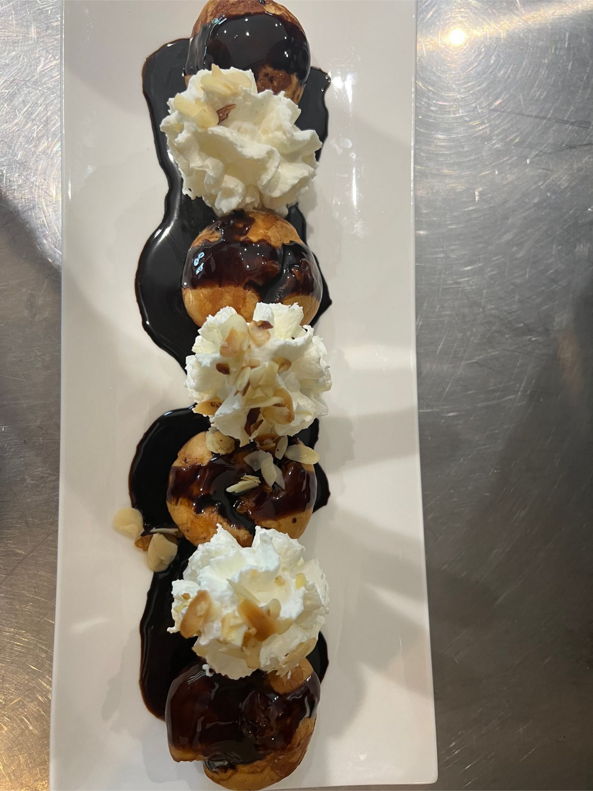 Profiteroles sur une assiette blanche, arrosées de sauce au chocolat et garnies de crème fouettée et d'amandes.