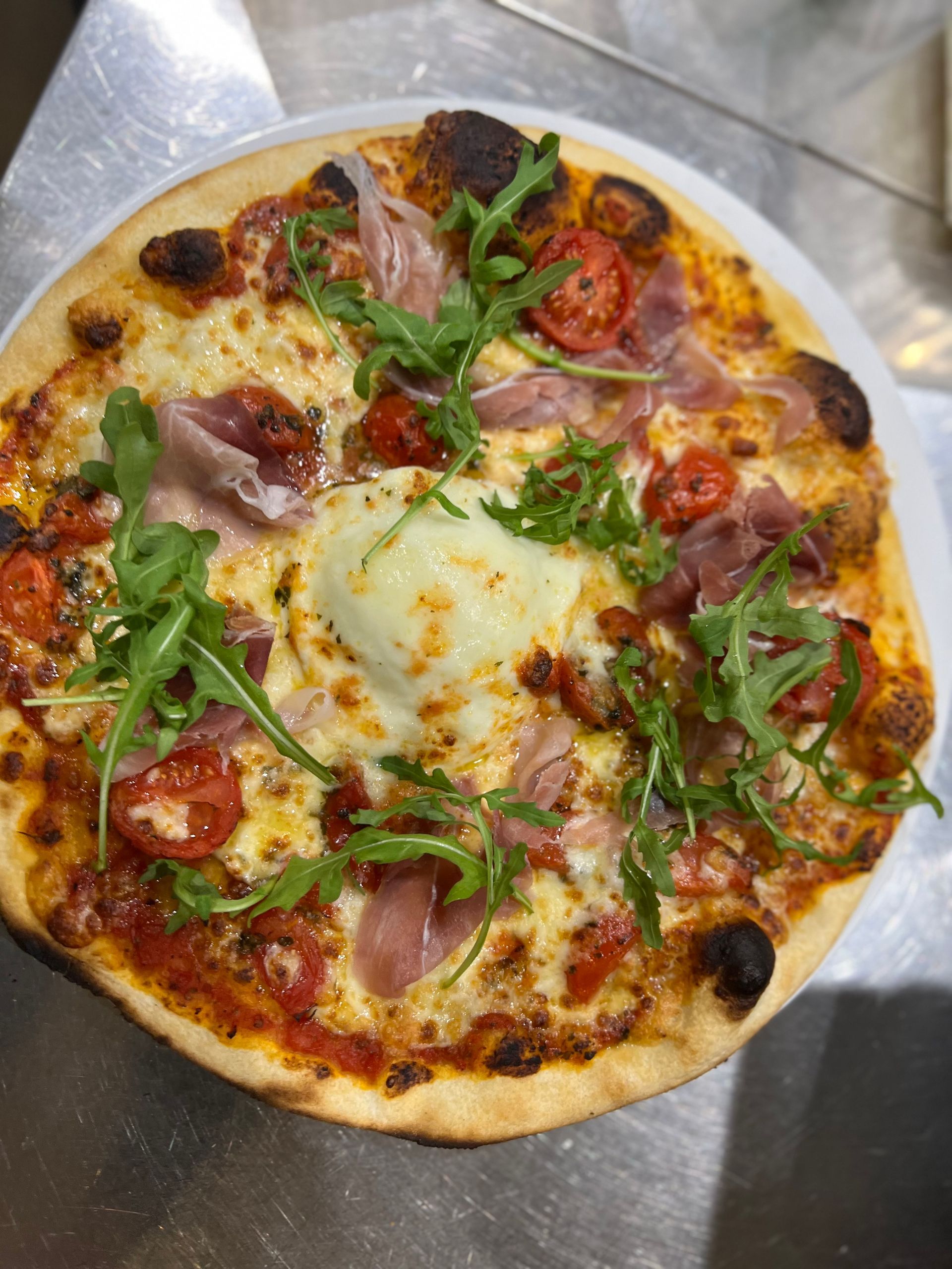 Pizza garnie de fromage, de prosciutto, de tomates et de roquette, servie dans une assiette blanche.