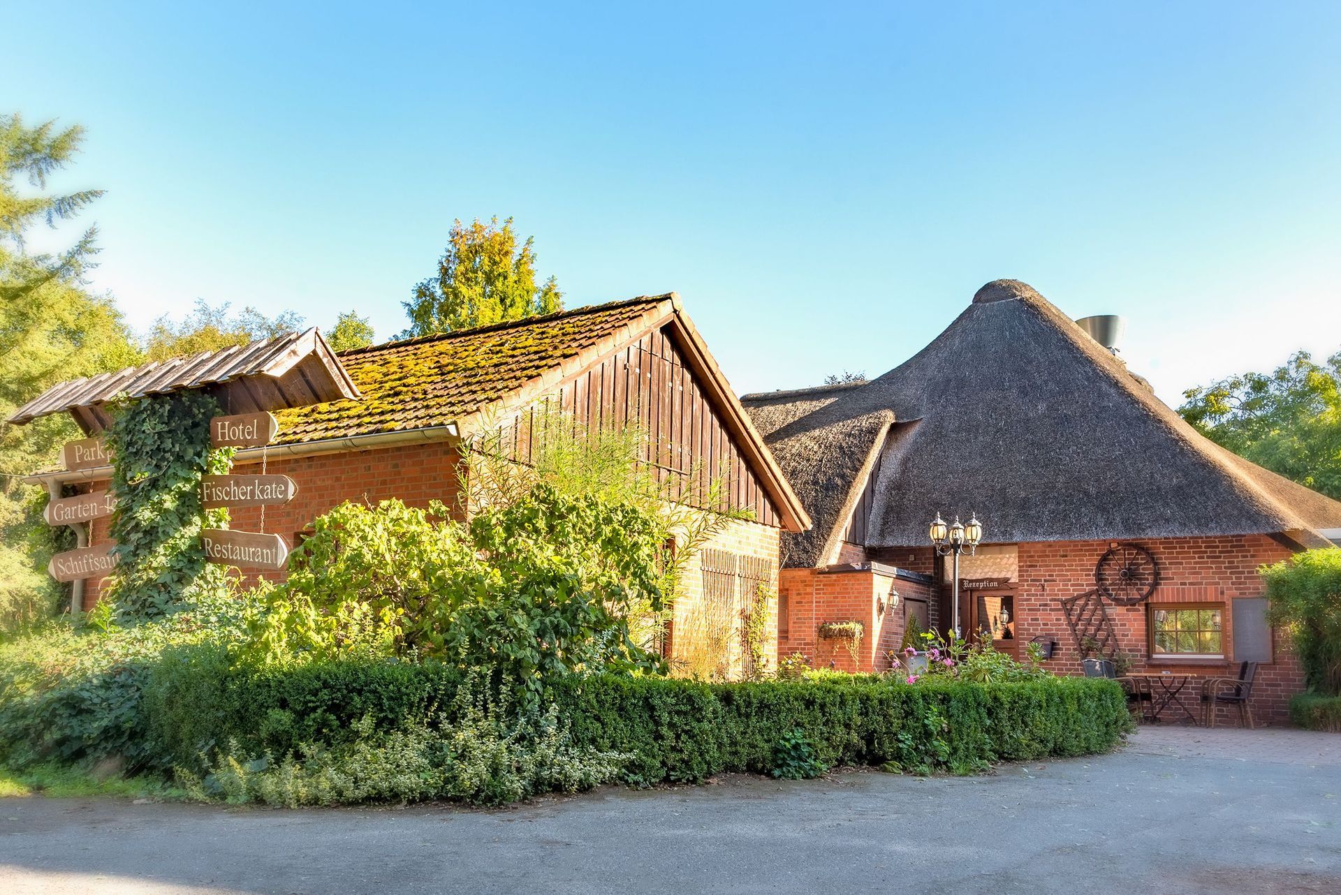 Ein rustikales Backsteingebäude mit Strohdach und hölzernen Wegweisern, eingebettet in eine grüne Landschaft unter blauem Himmel.