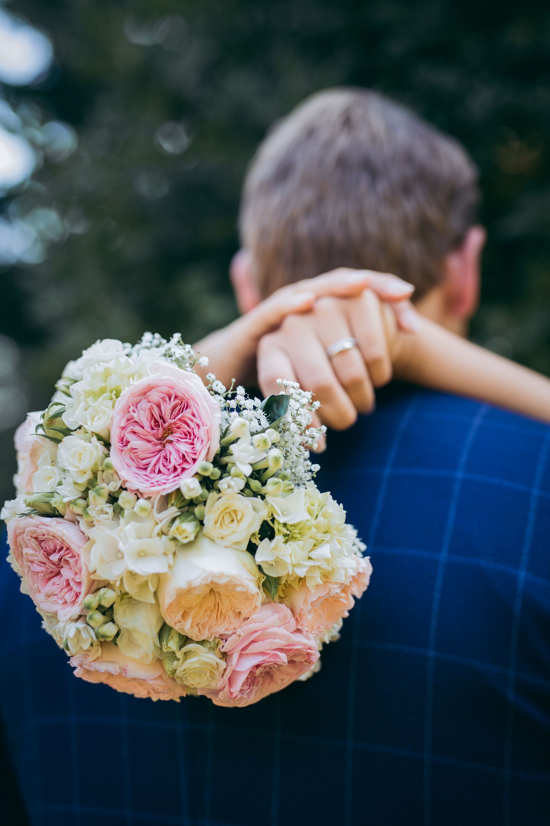 Die Arme einer Frau liegen um den Hals eines Mannes, ein Ring ist sichtbar. Sie hält einen Strauß rosa und weißer Blumen im Freien.