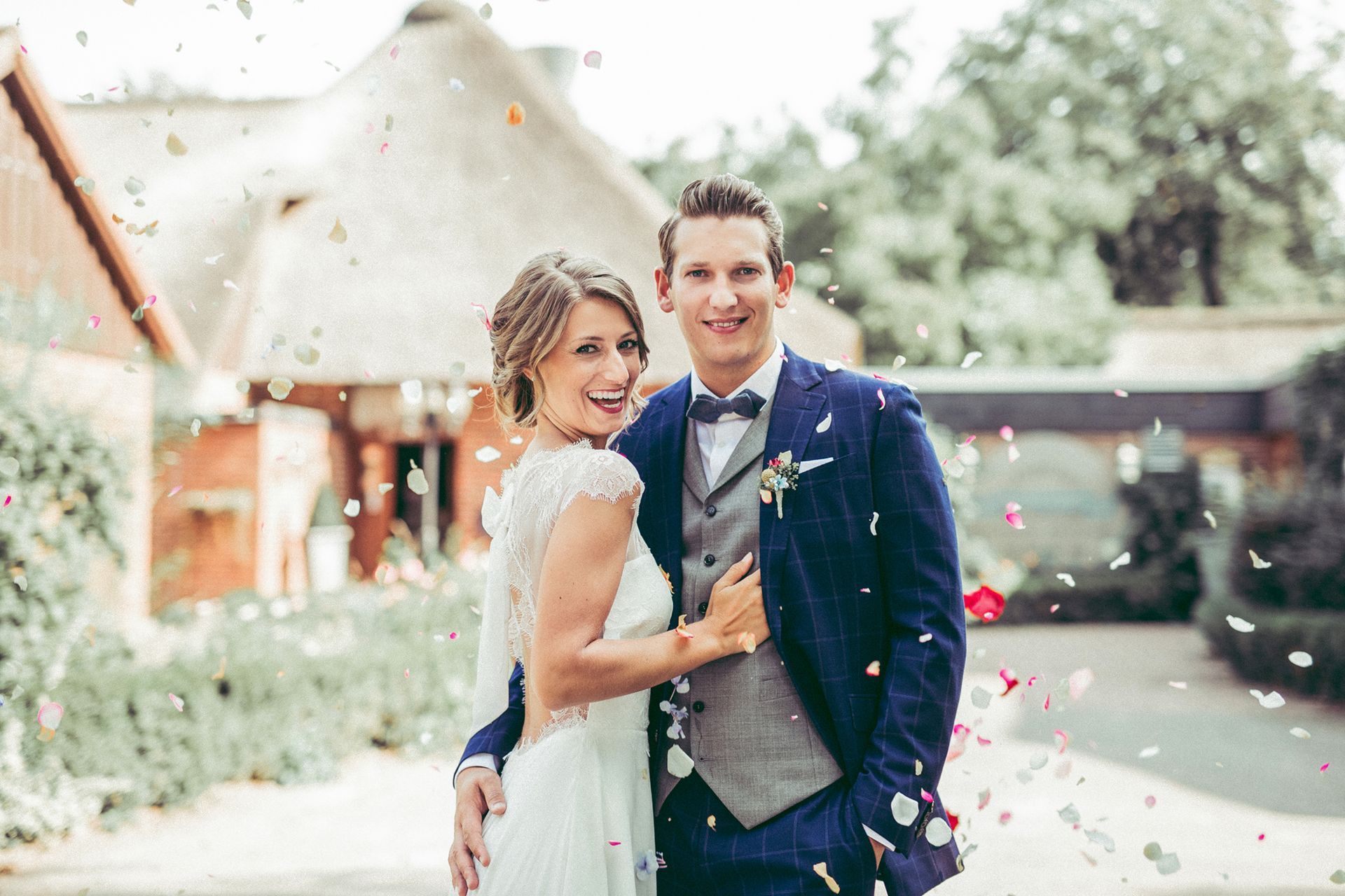 Braut und Bräutigam lächeln, Konfetti fällt vor einem Gebäude mit Strohdach.