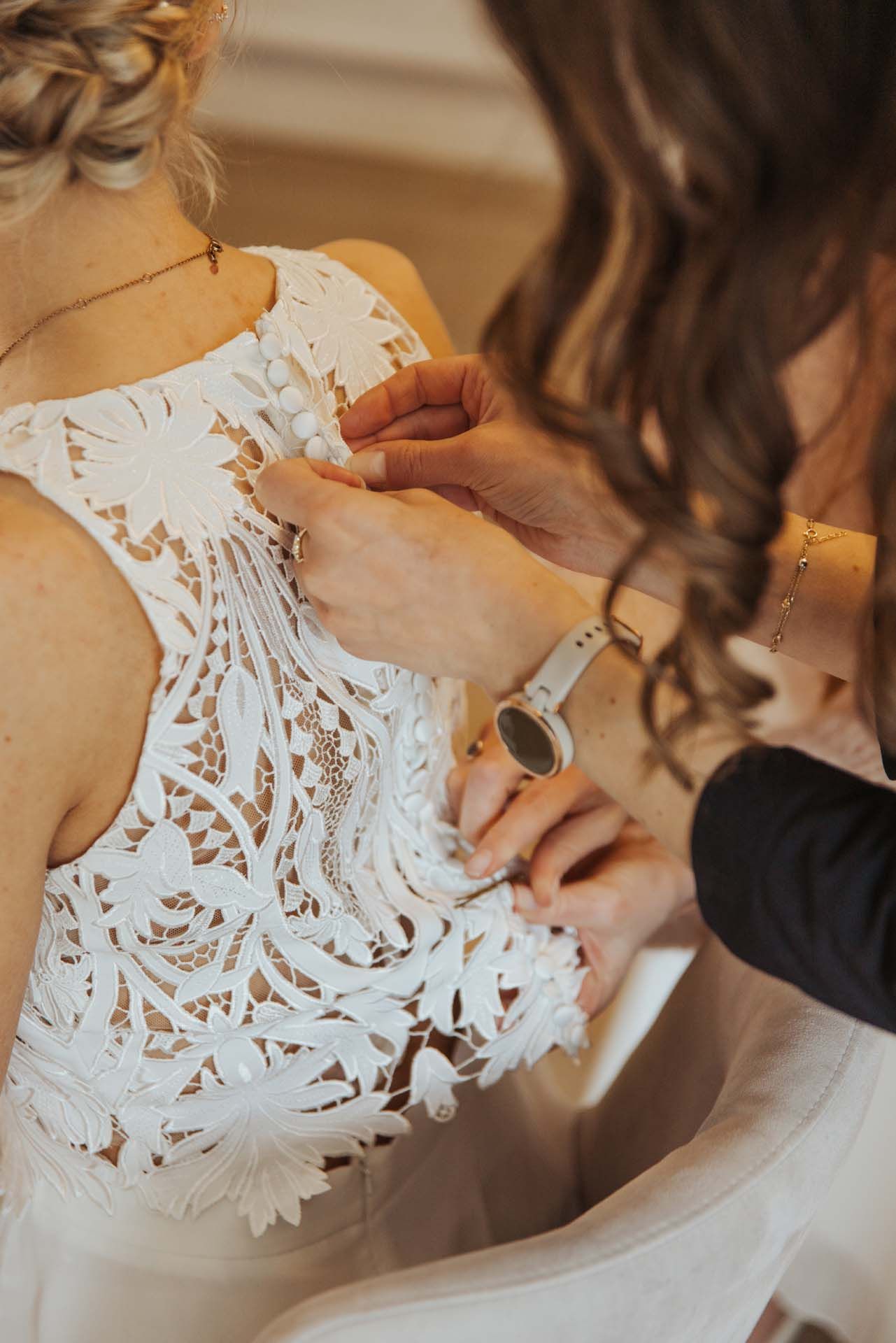 Der Rücken einer Frau in einem weißen Spitzenkleid wird von einer anderen Person zugeknöpft, wahrscheinlich einer Braut, die sich fertig macht.