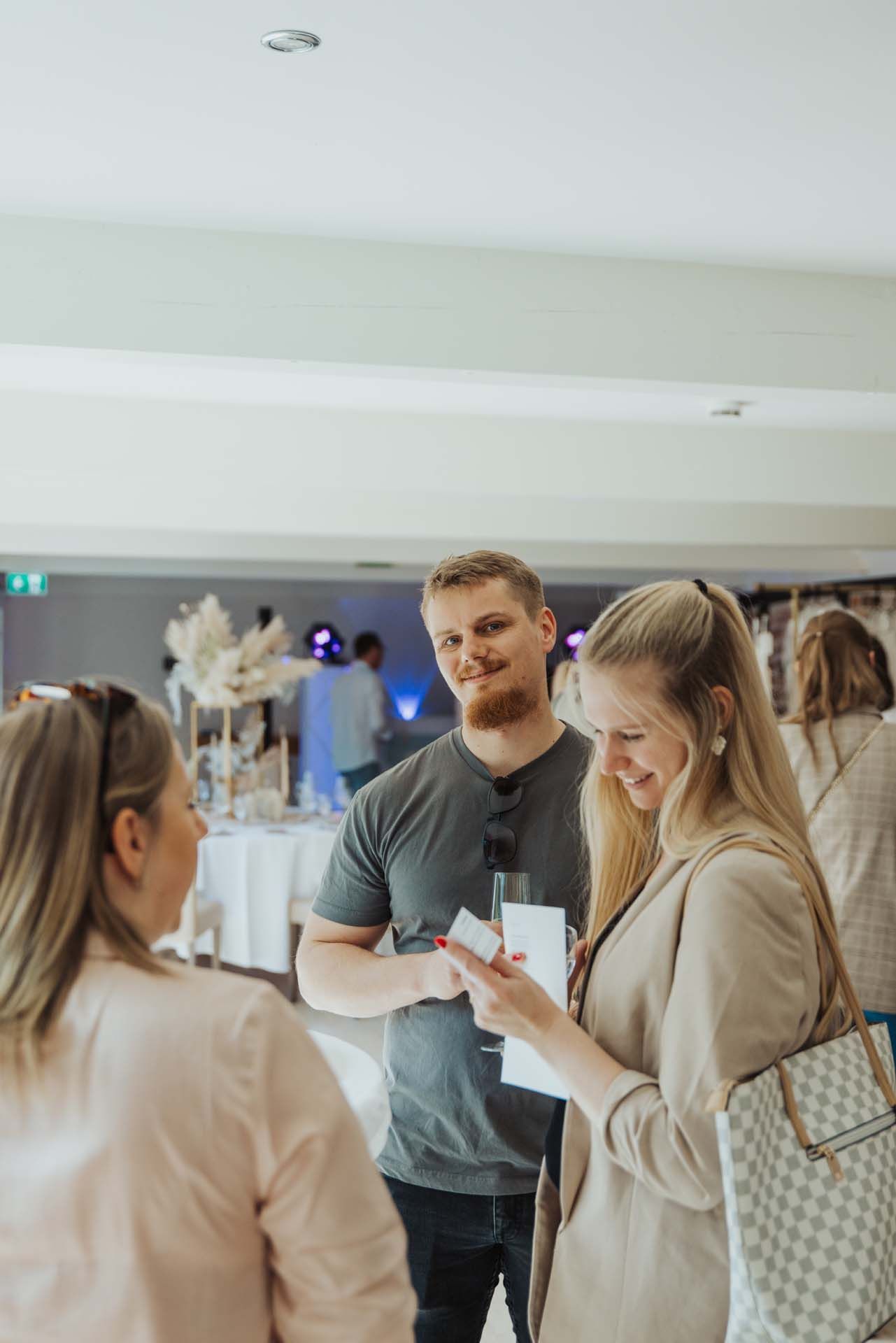 Drei Personen unterhalten sich bei einer Hochzeitsfeier: ein Mann, zwei Frauen. Eine Frau zeigt ihr Telefon; alle lächeln. Weißes Ambiente.