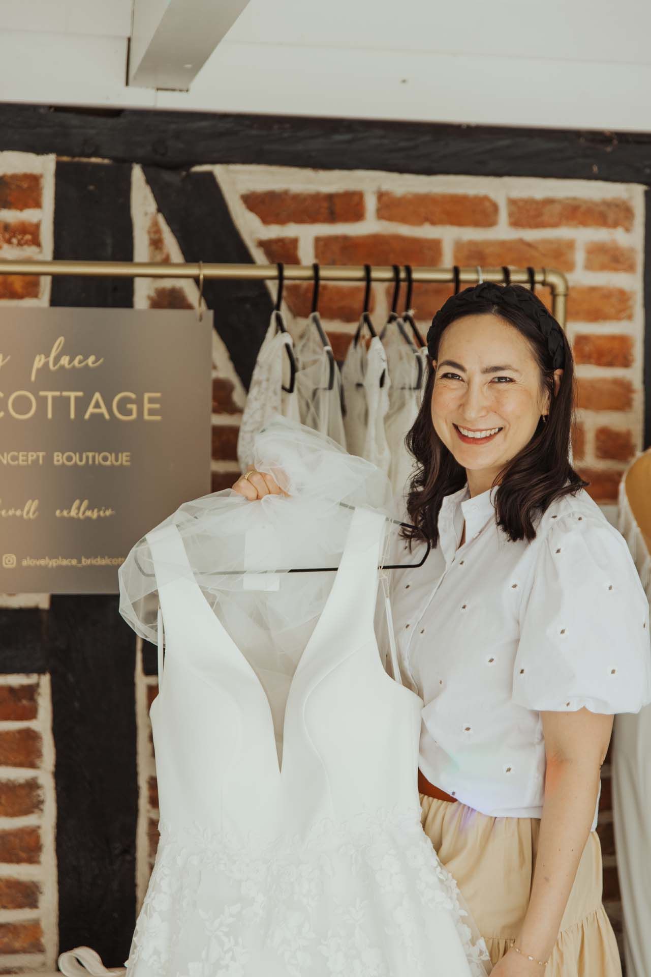 Frau lächelt und hält ein weißes Hochzeitskleid. Boutique-Einstellung, Backsteinmauer-Hintergrund.