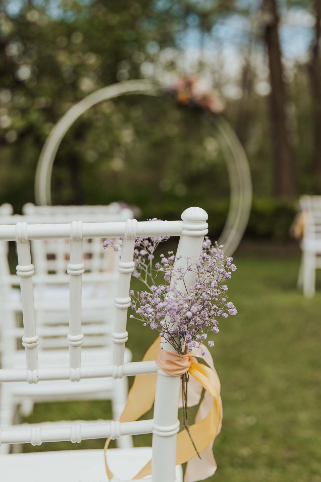 Weiße Stühle mit Blumendekorationen, gedeckt für eine Zeremonie im Freien, mit einem Blumenbogen im Hintergrund.