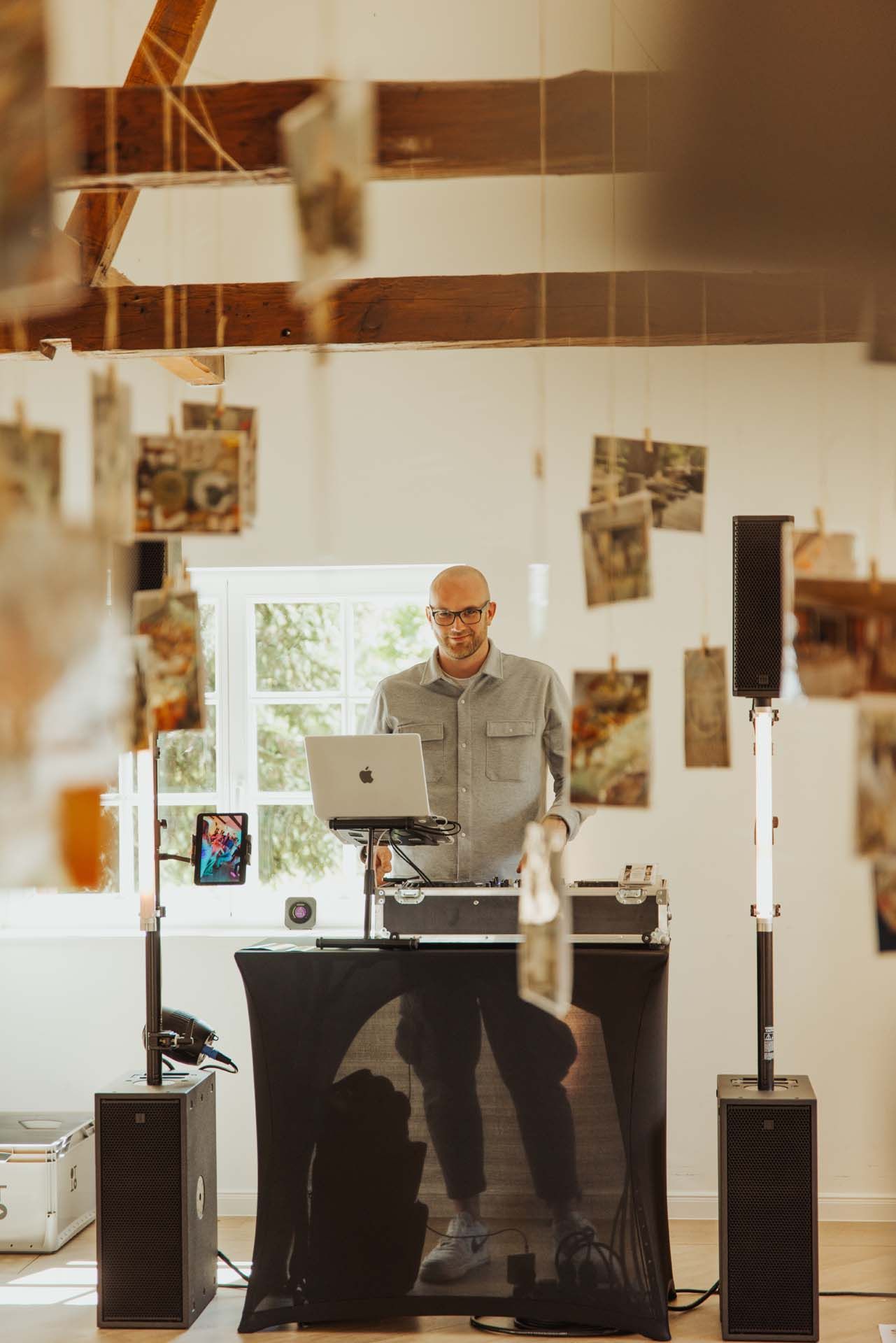 DJ in einem weißen Raum, hinter einer schwarzen Kabine mit Lautsprechern stehend. Sonnenlicht strömt herein, Fotos hängen.
