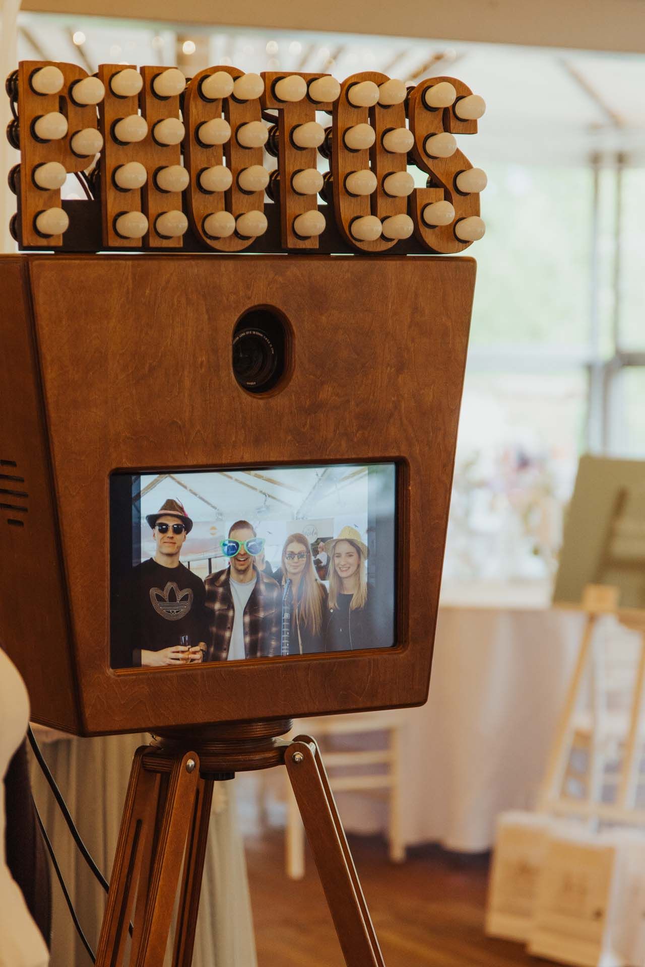 Fotoautomat aus Holz mit „KISS“-Schriftzug, der ein Gruppenfoto anzeigt. Dreibeinständer in heller Umgebung.