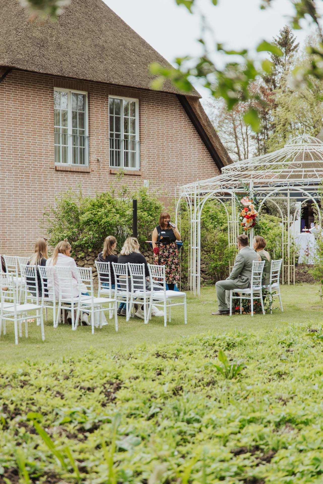Hochzeitszeremonie im Freien: Gäste sitzen, der Geistliche spricht neben dem Pavillon, im Hintergrund ein Backsteingebäude.