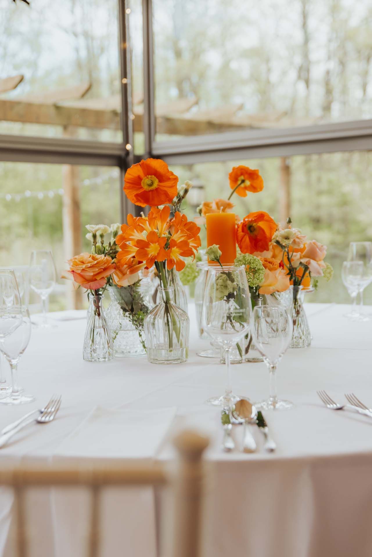 Tafelaufsatz aus orangefarbenen Blumen in Glasvasen auf einer weißen Tischdecke. Esstisch in der Nähe eines großen Fensters.