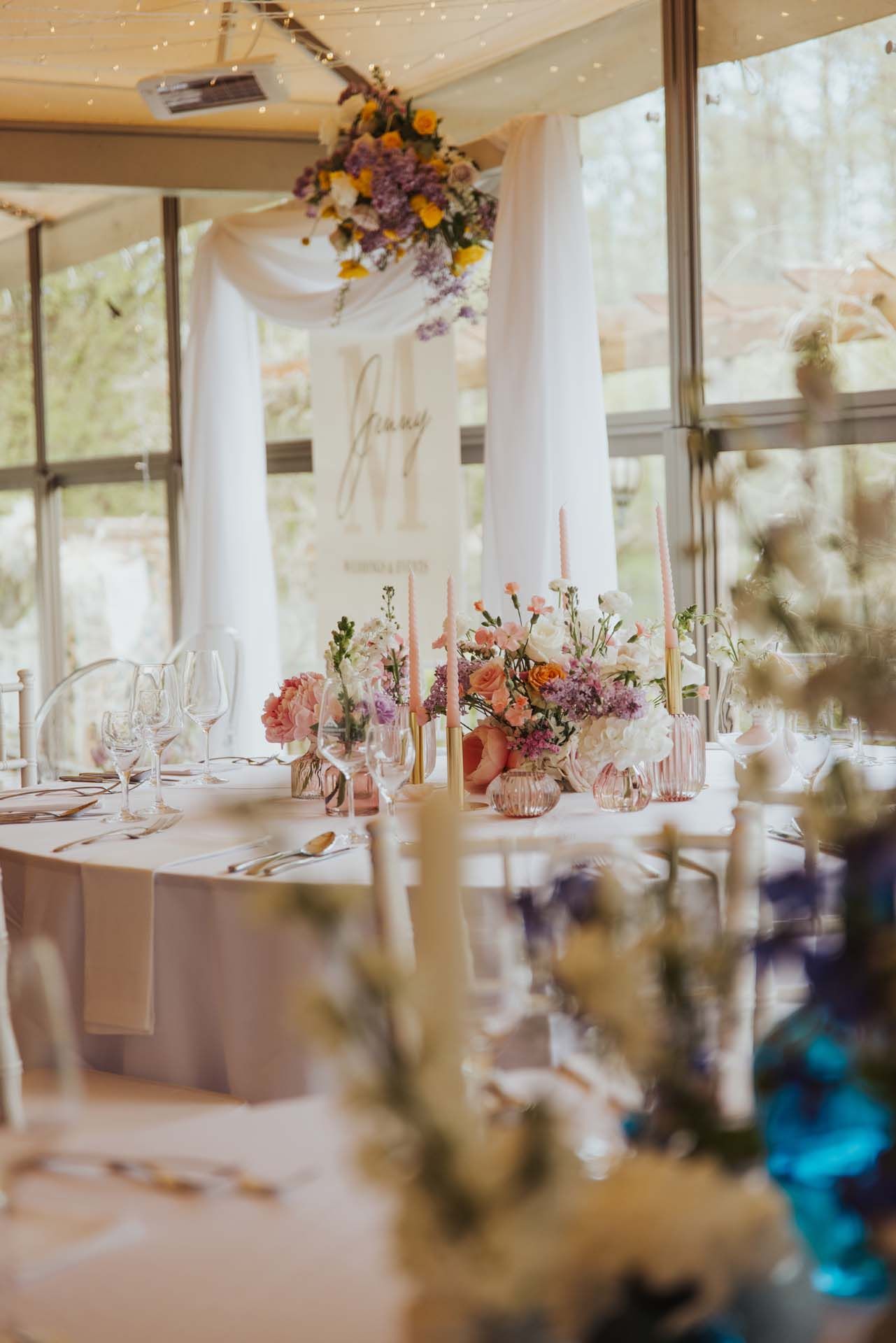 Hochzeitsempfangstisch mit Blumendekoration und drapiertem Bogen vor einer Glasfensterkulisse.