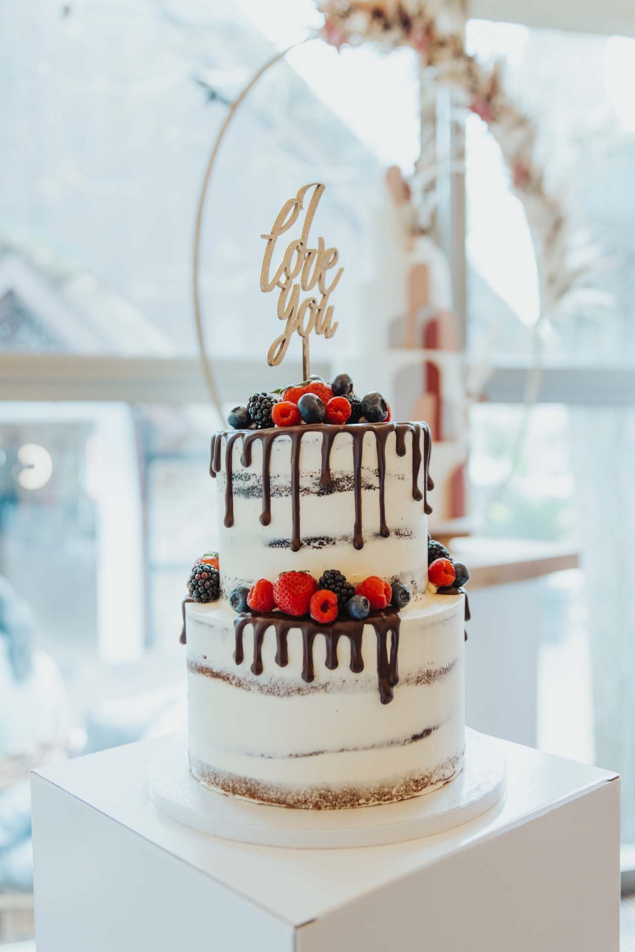 Zweistöckiger Kuchen mit Schokoladentropfen und Beeren, gekrönt mit einem „Ich liebe dich“-Schild auf einem weißen Ständer.