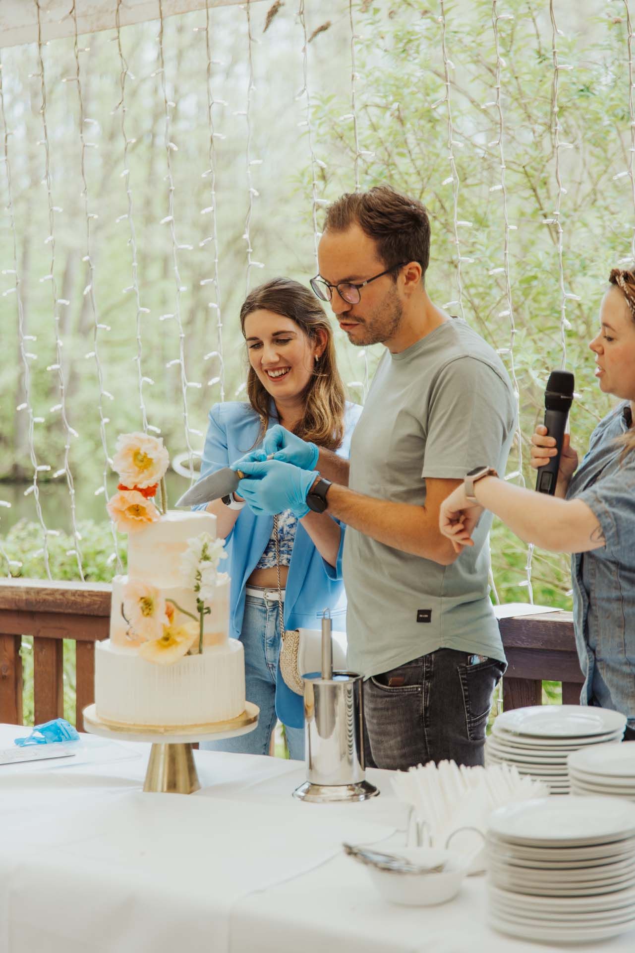 Paar schneidet im Freien eine Hochzeitstorte an; Frau lächelt, Mann trägt Handschuhe, Person hält Mikrofon.