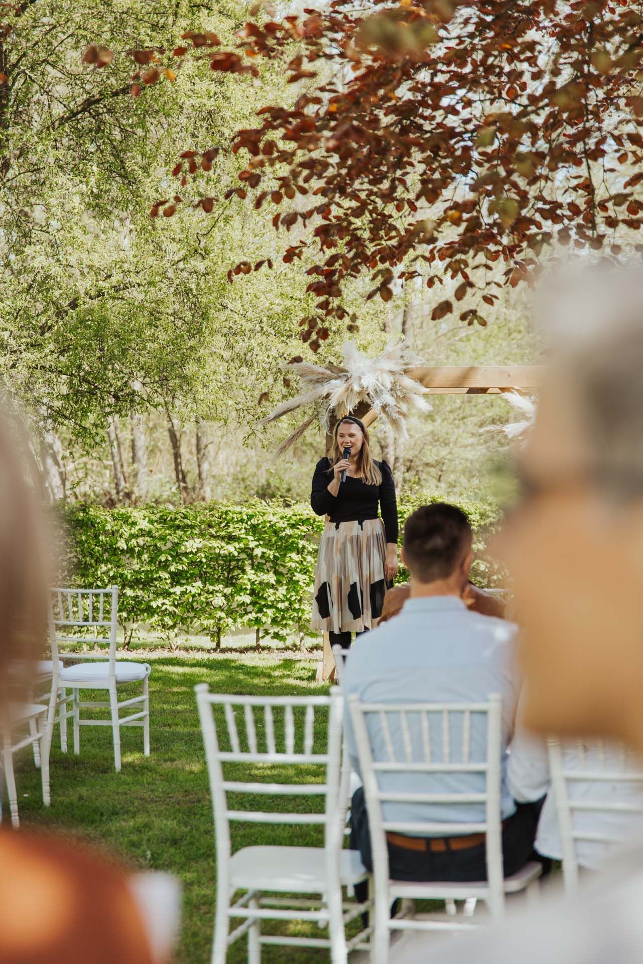 Frau spricht bei einer Hochzeitszeremonie im Freien. Gäste sitzen auf weißen Stühlen, im Hintergrund Grün.