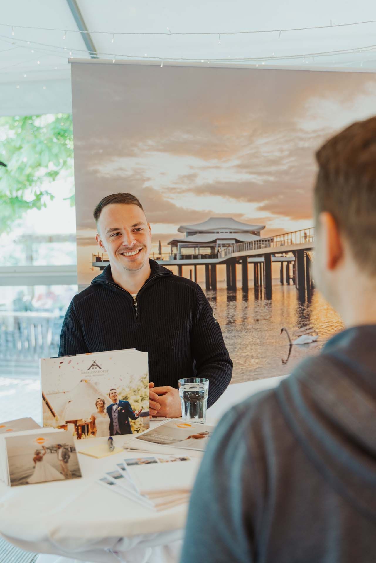 Ein Mann lächelt einen Kunden an und sitzt an einem Tisch mit Broschüren und Fotos; im Hintergrund ein Pier bei Sonnenuntergang.