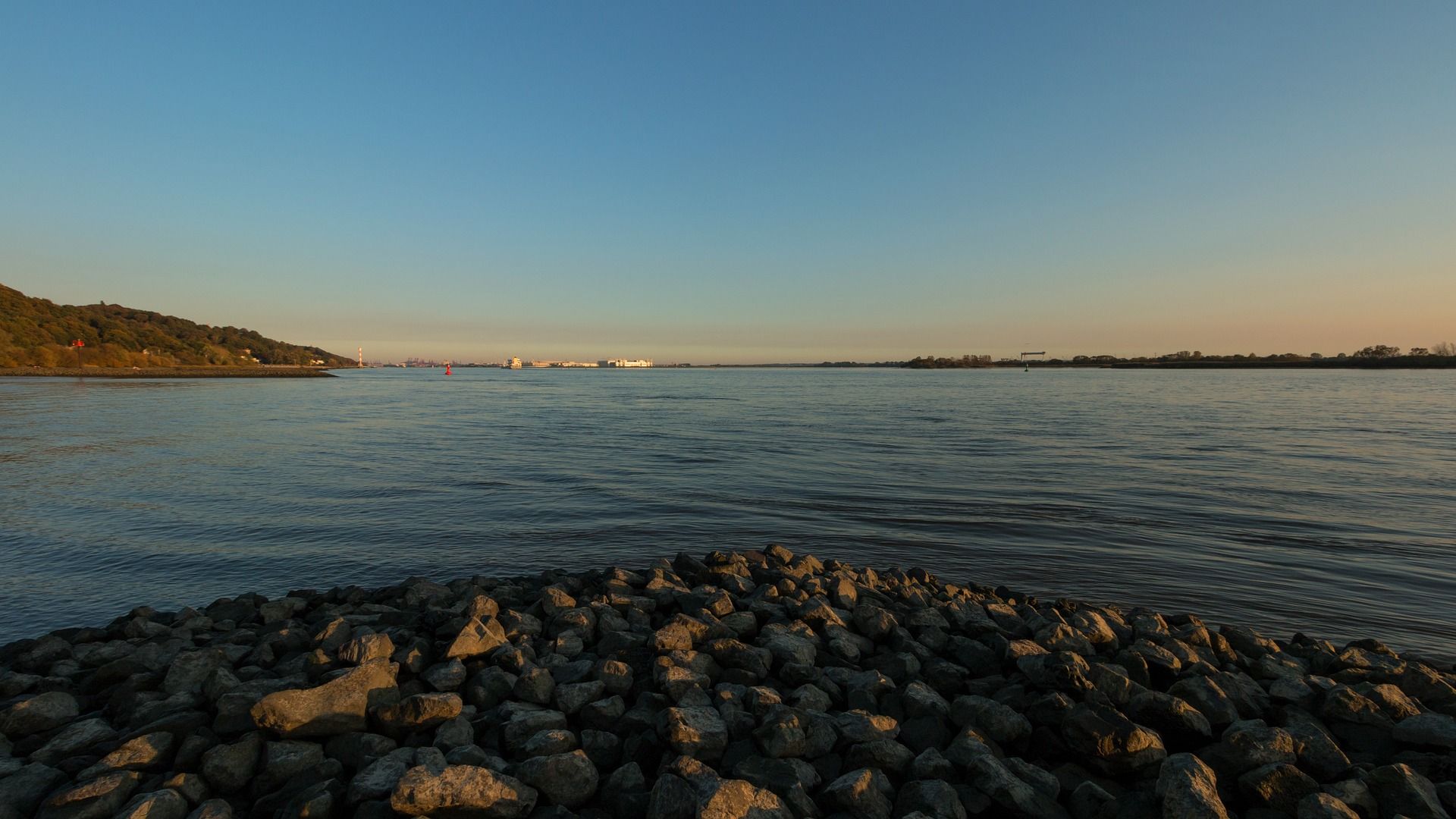 Ruhiges Wasser mit felsigem Ufer unter einem klaren blauen Himmel, Gebäude in der Ferne.