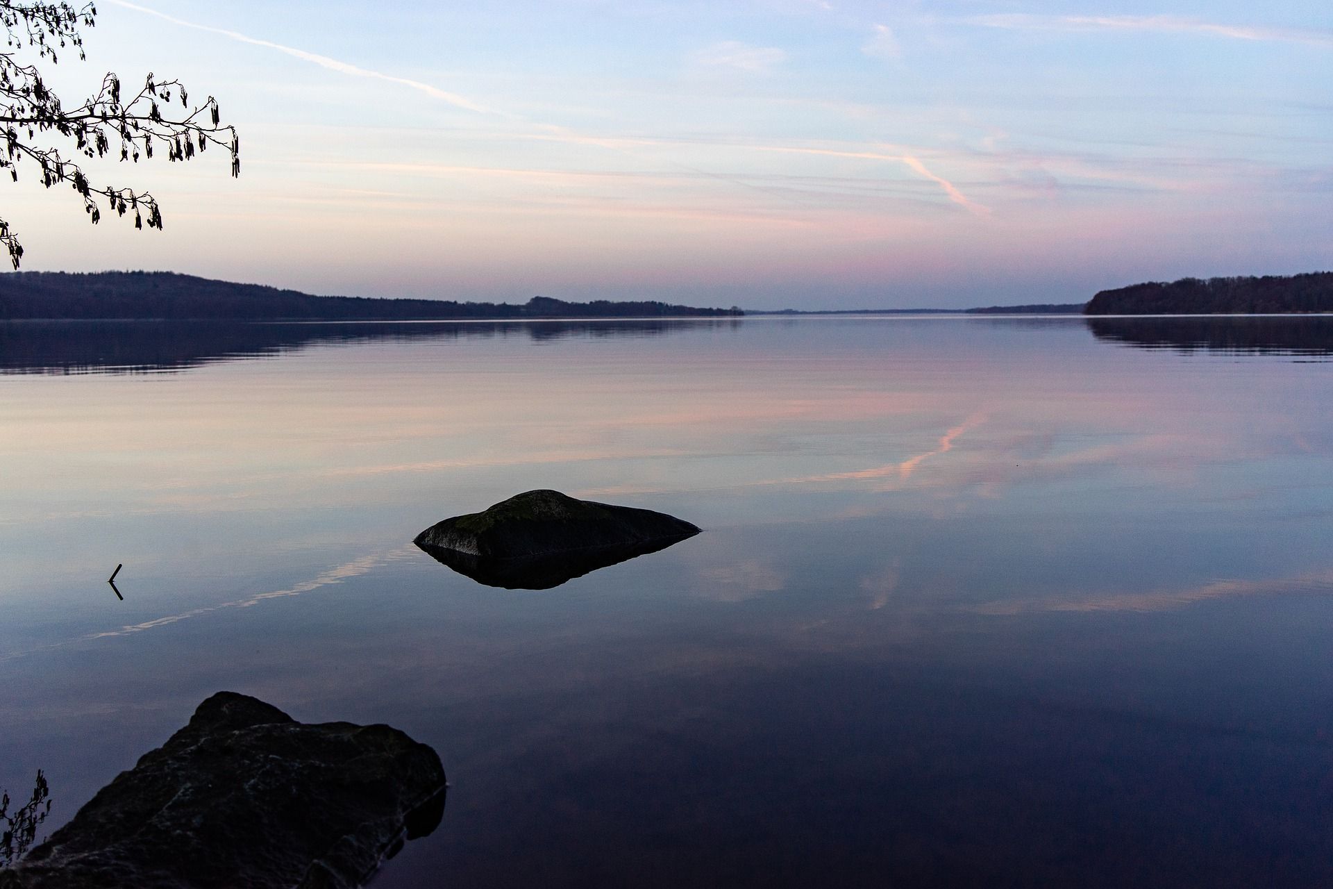 Ruhiger See in der Abenddämmerung, der den zartrosa und blauen Himmel reflektiert. Eine Felseninsel liegt mitten im Wasser.