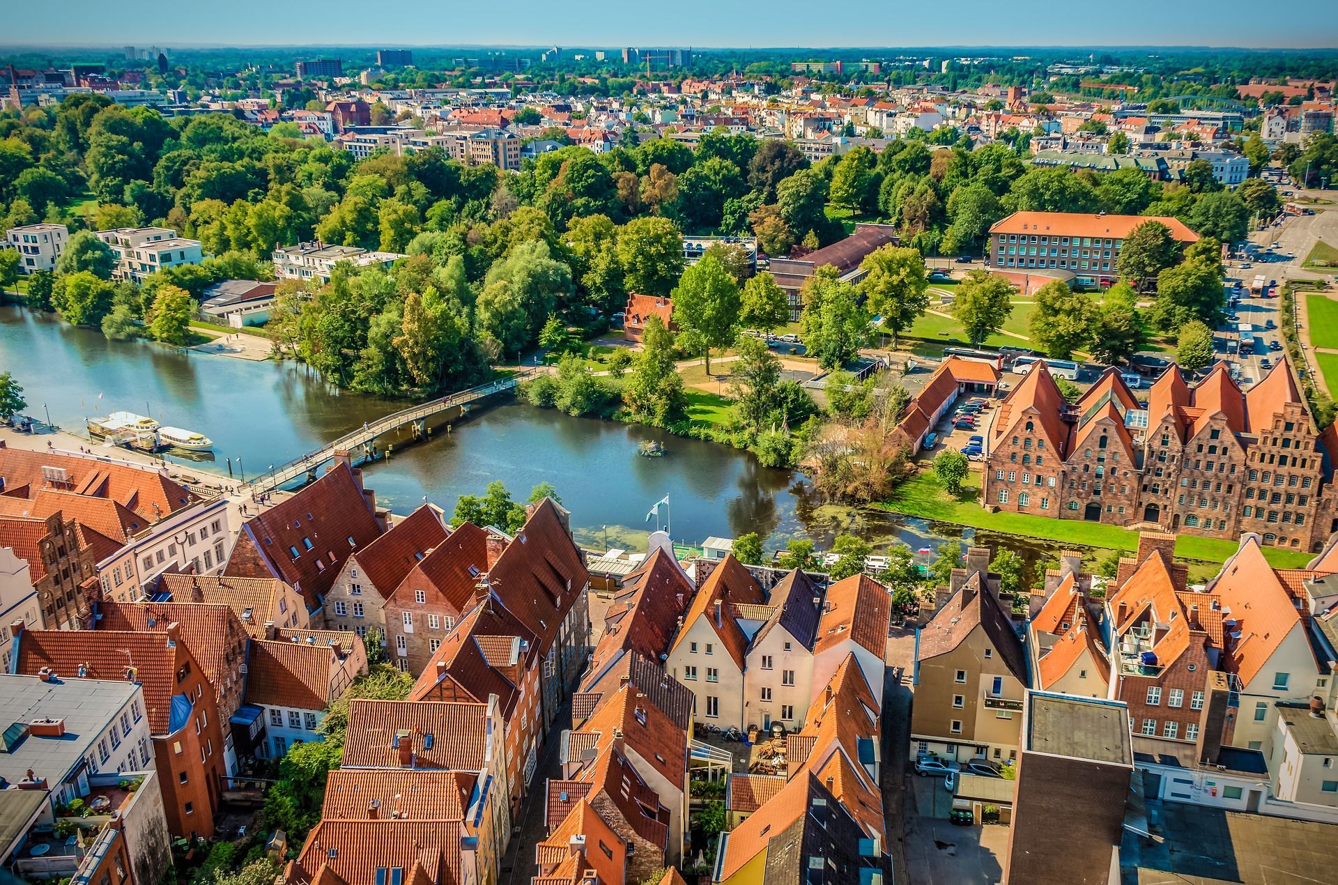 Luftaufnahme von Lüneburg, Deutschland, mit Gebäuden mit roten Dächern, einem Fluss, einer Brücke und üppig grünen Bäumen unter einem klaren blauen Himmel.