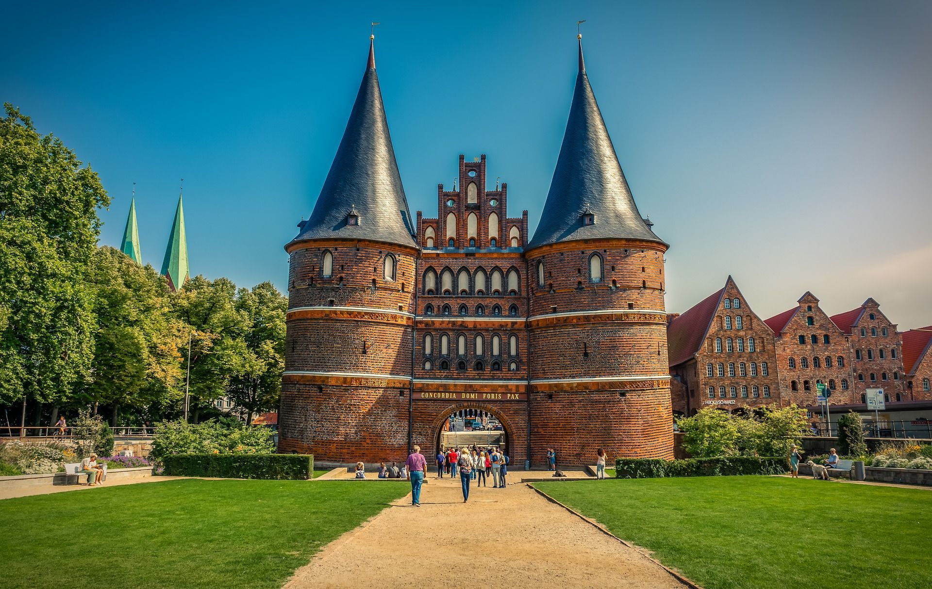 Holstentor in Lübeck, Deutschland, mit zwei Türmen, blauen kegelförmigen Dächern und Menschen, die auf dem Weg spazieren.