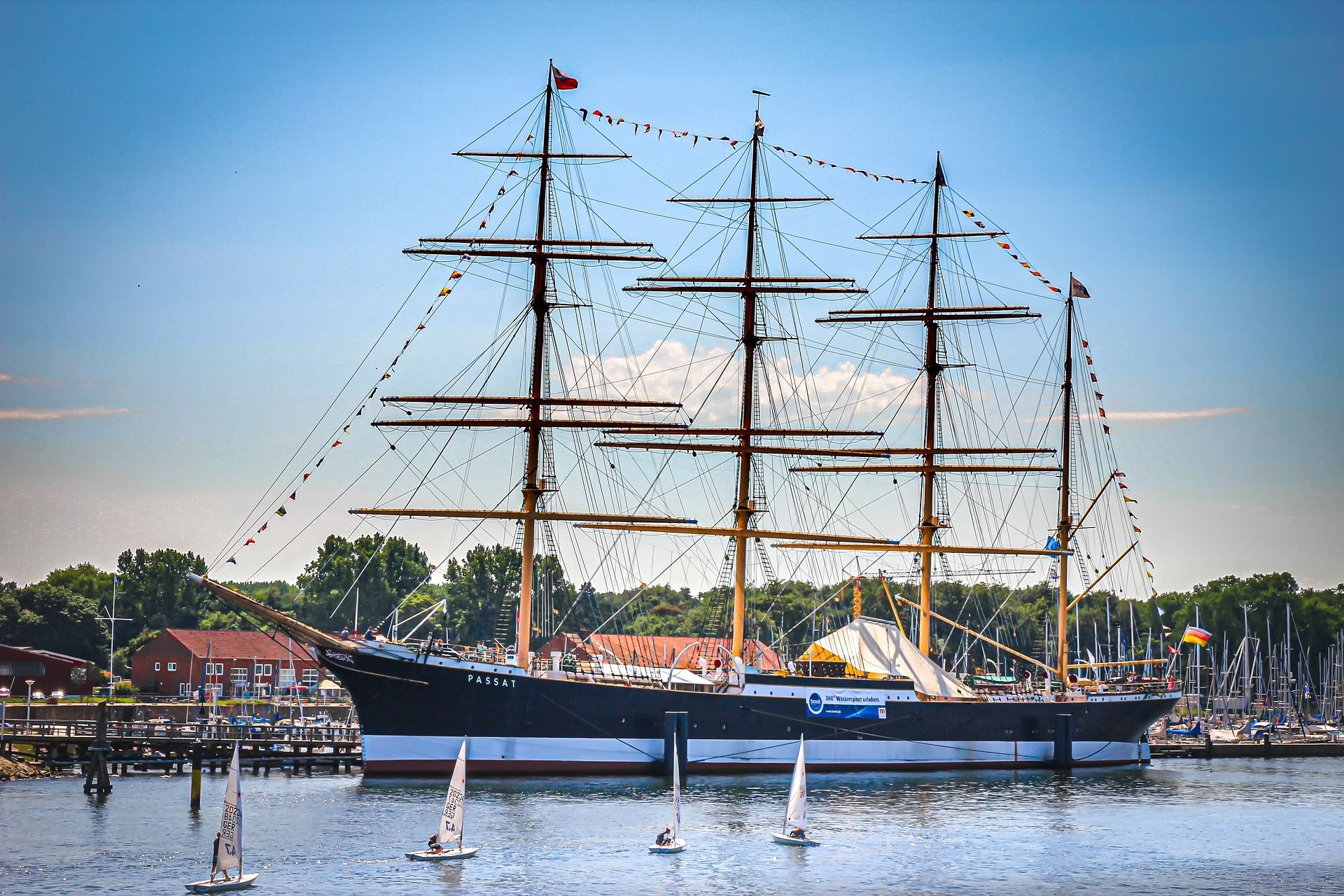 Großes Schiff im Hafen, schwarzer Rumpf, weiße Streifen, mehrere Masten, blauer Himmel, kleine Segelboote im Wasser.
