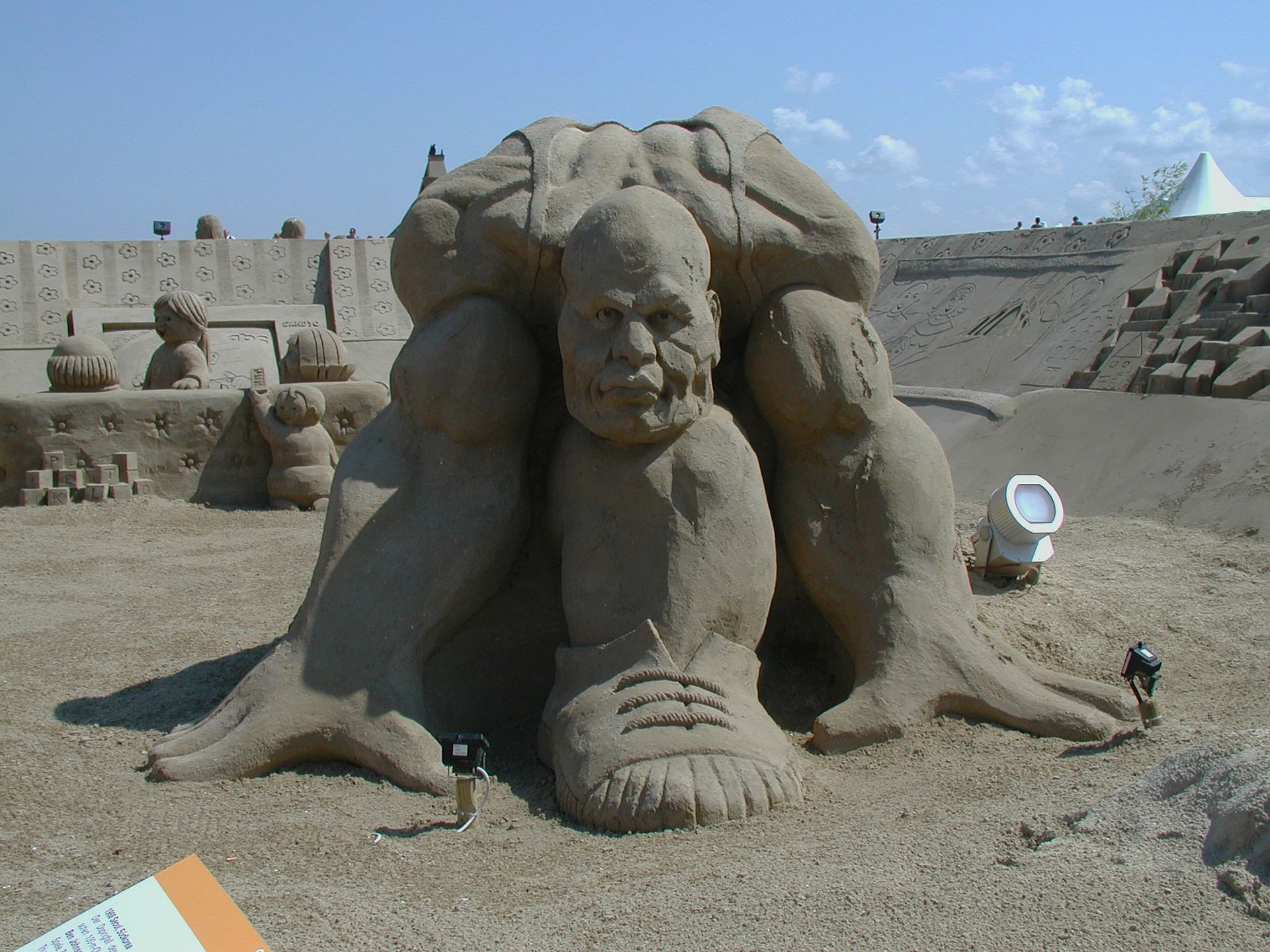 Sandskulptur einer menschlichen Figur mit Armen und Beinen und detaillierten Gesichtszügen. Im Freien, unter blauem Himmel.