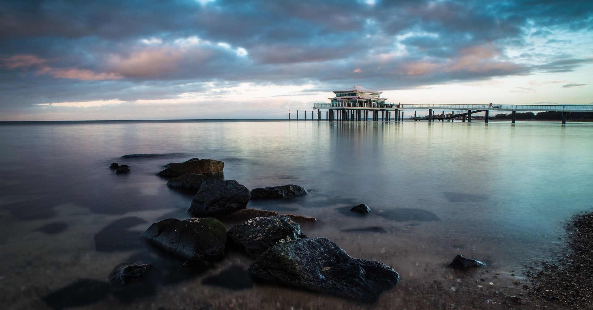 Ruhiges Wasser in der Abenddämmerung mit einem Pier in der Ferne, Felsen im Vordergrund und bewölktem Himmel.