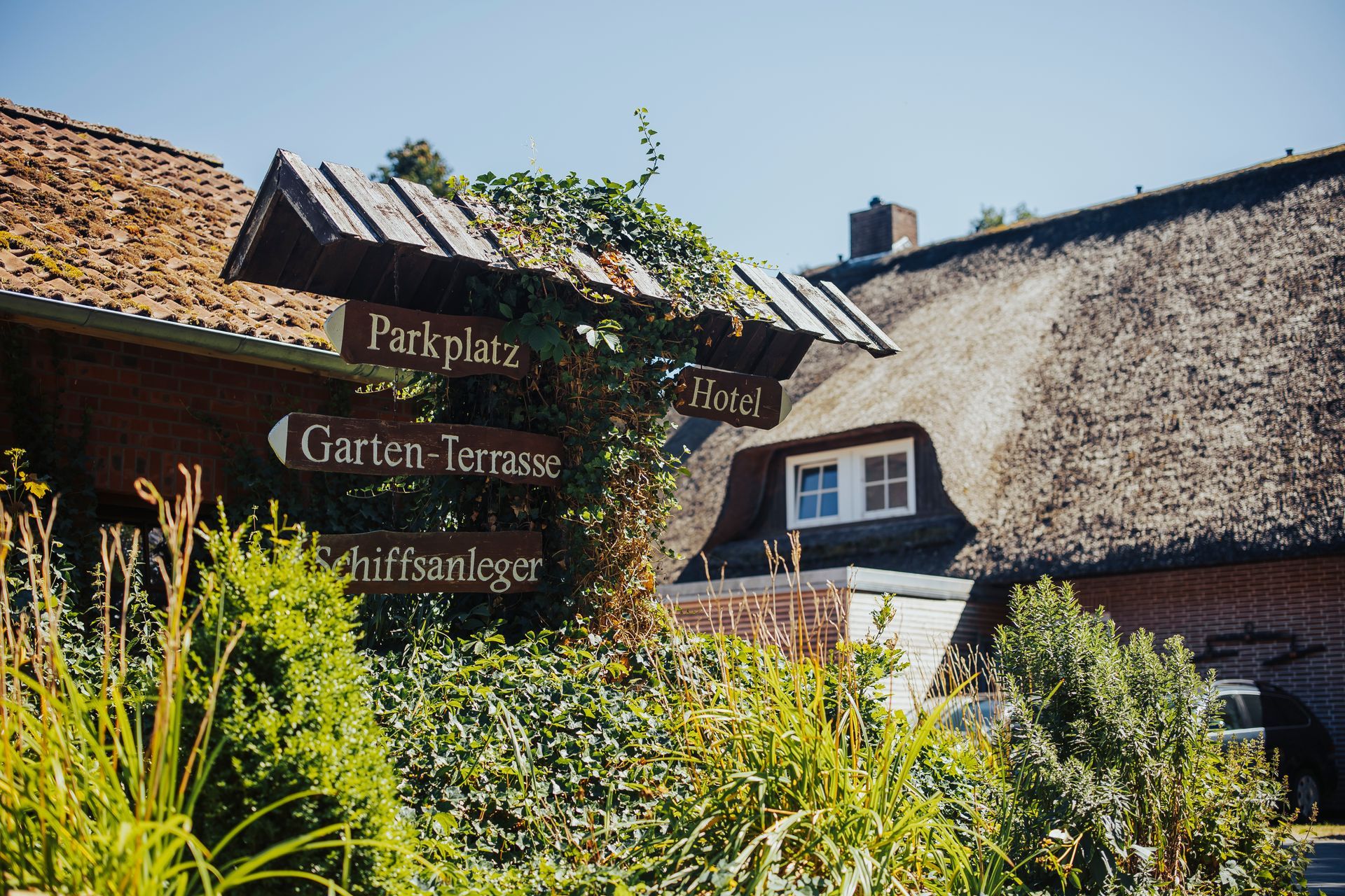 Holzwegweiser mit Wegbeschreibung: Parkplatz, Gartenterrasse und Hotel, neben einem Gebäude mit Strohdach.