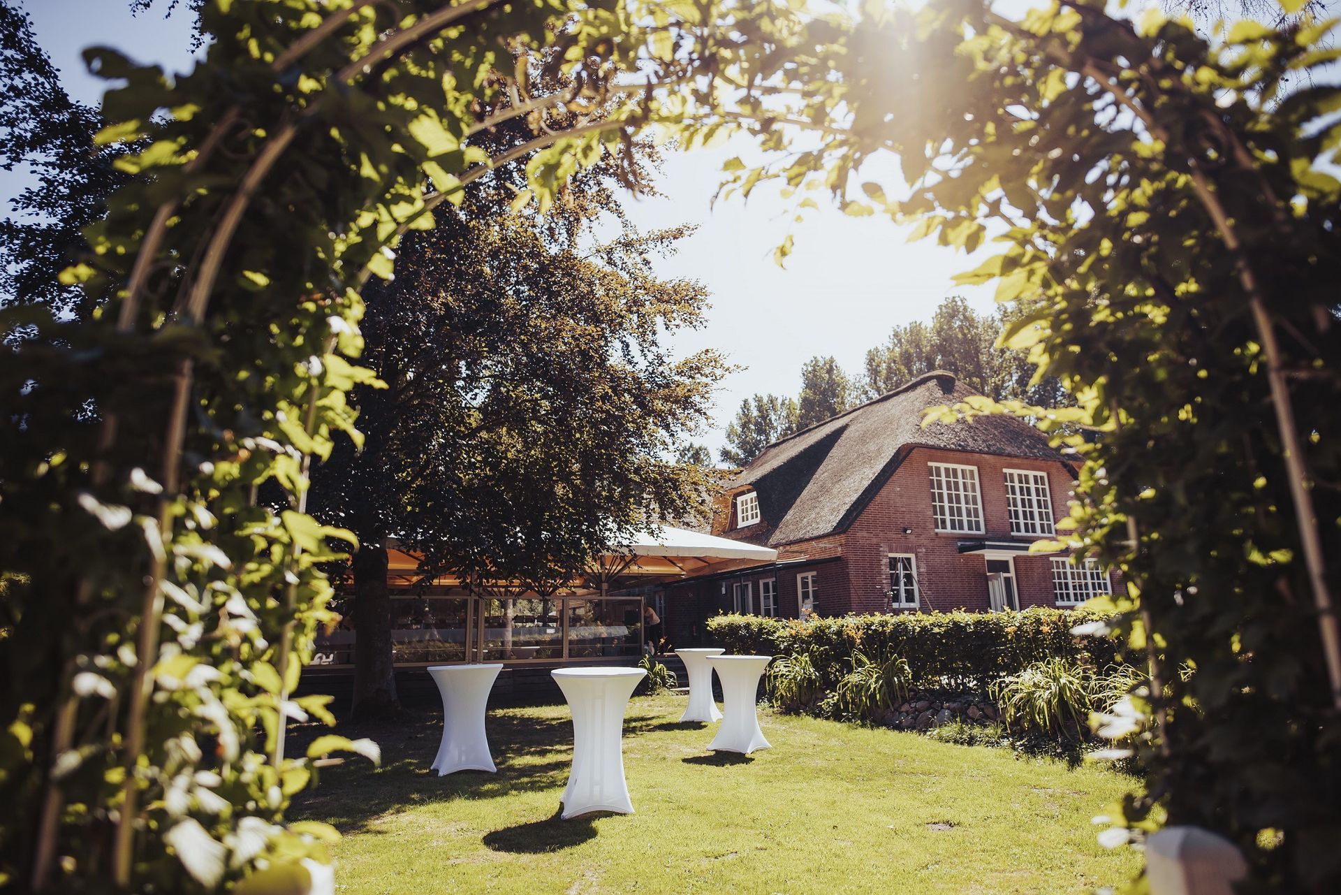 Gewölbtes Laub umrahmt eine sonnige Gartenszene mit einem Backsteingebäude und weißen Cocktailtischen.
