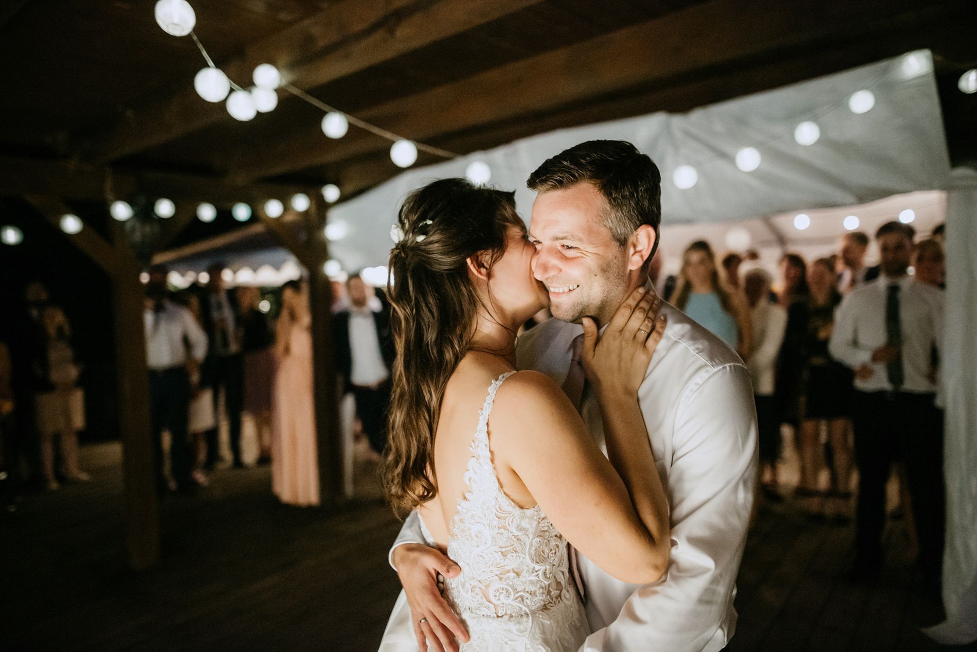 Die Braut küsst den Bräutigam während eines Tanzes bei einem Hochzeitsempfang im Freien auf die Wange; Lichterketten und Gäste im Hintergrund.