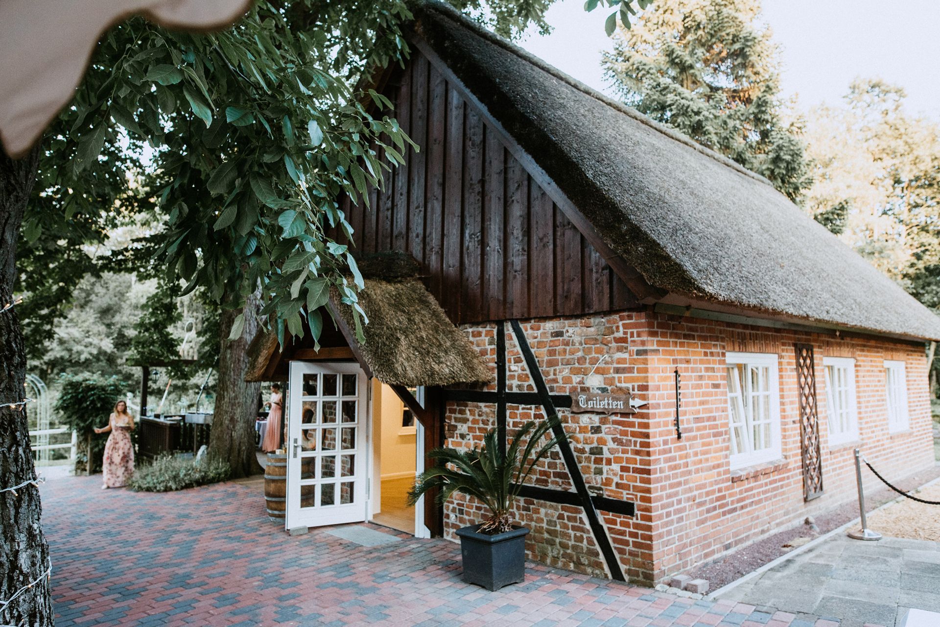Backsteinhaus mit Strohdach, weißer Tür und weißen Fenstern. Bäume umgeben das Gebäude in einer parkähnlichen Umgebung.