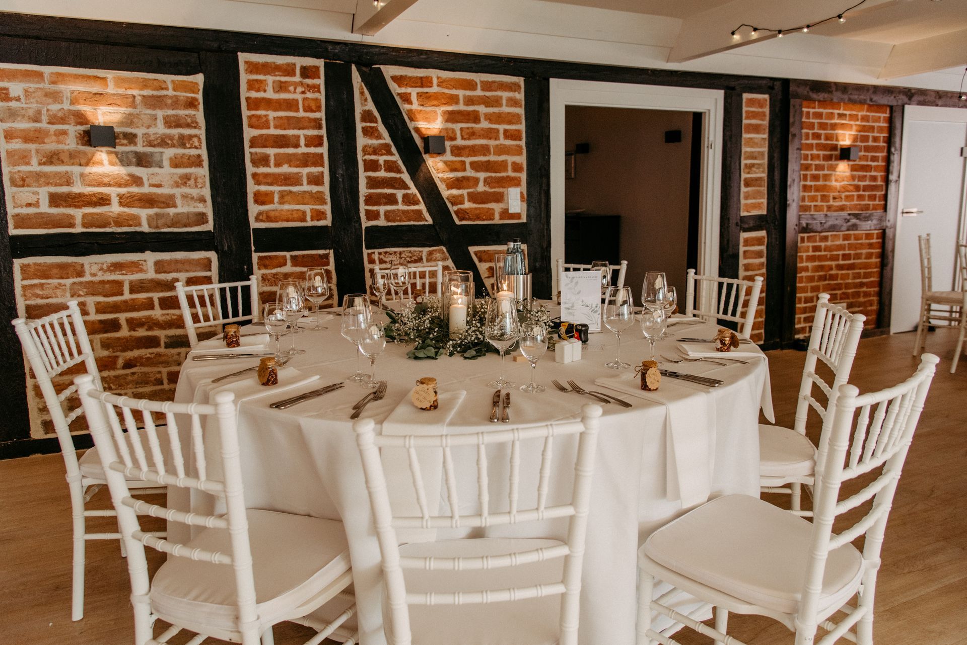 Runder Tisch für eine Feier mit weißen Stühlen, einer Ziegelwand im Hintergrund und Grünpflanzen in der Mitte.