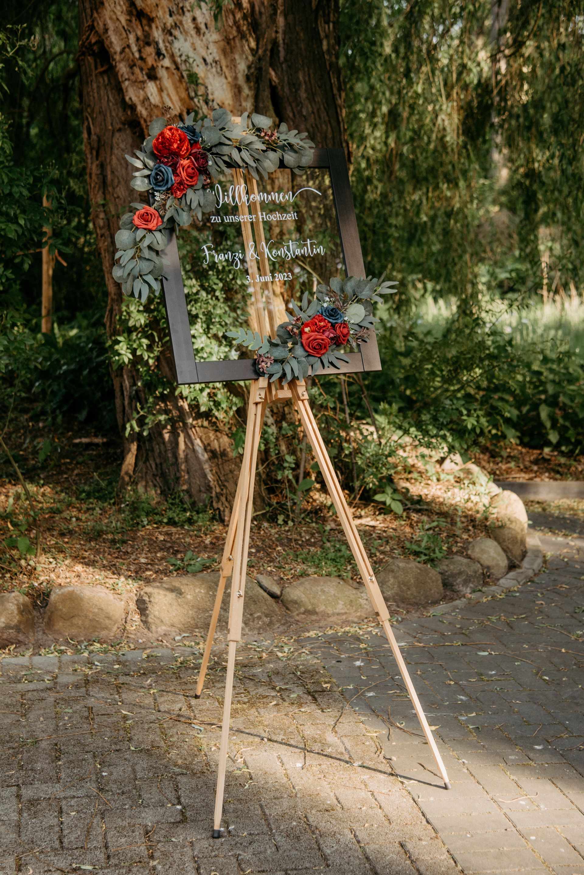 Hölzerne Staffelei mit einem klaren Schild, grau gerahmt und mit Blumen geschmückt, im Freien.