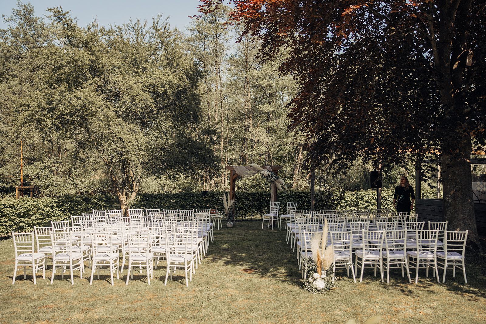 Hochzeitszeremonie mit weißen Stühlen auf Gras unter Bäumen.