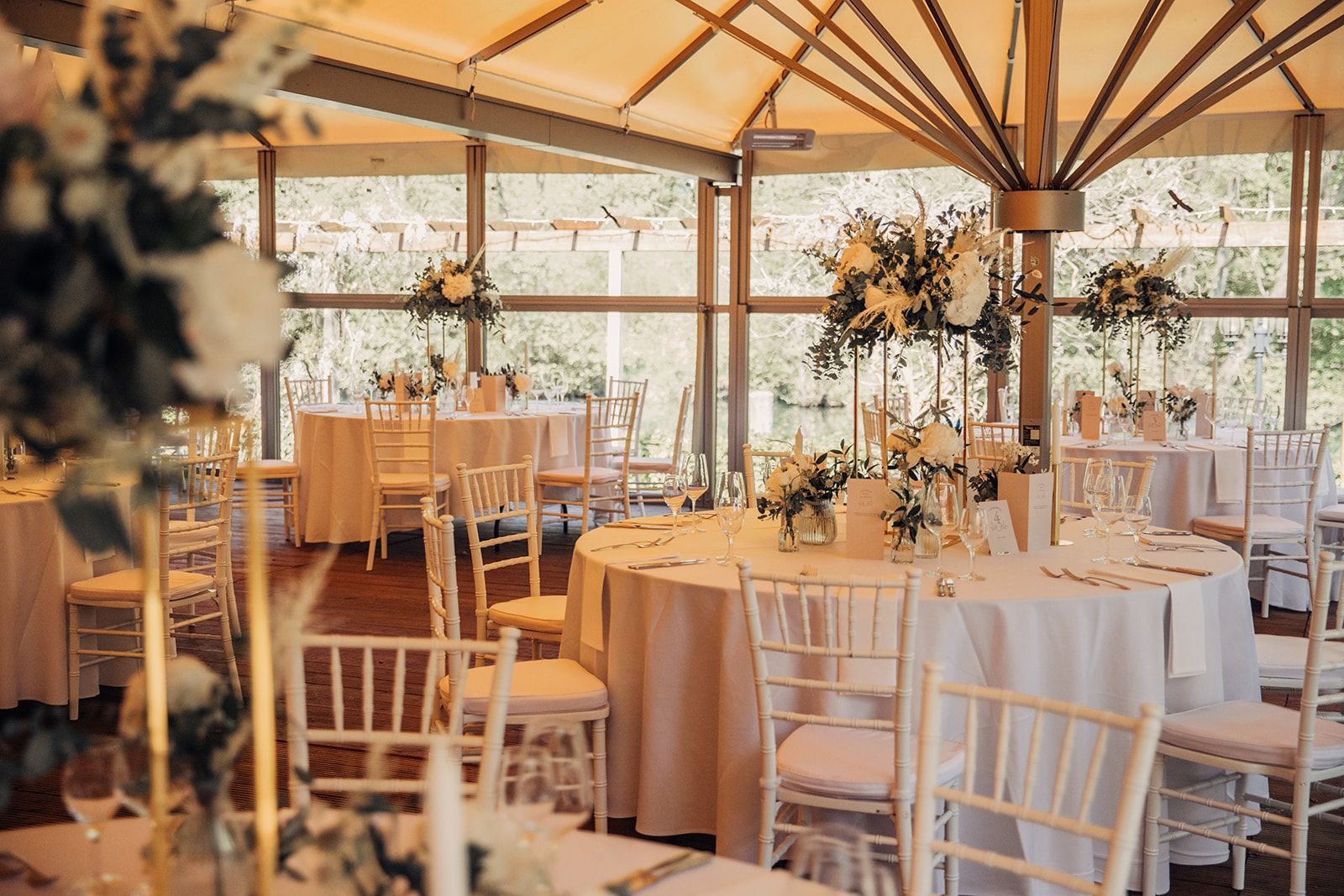 Eleganter Hochzeitsempfang, runde Tische mit weißer Tischwäsche und Blumengestecken in einem Veranstaltungsort mit Glaswänden.
